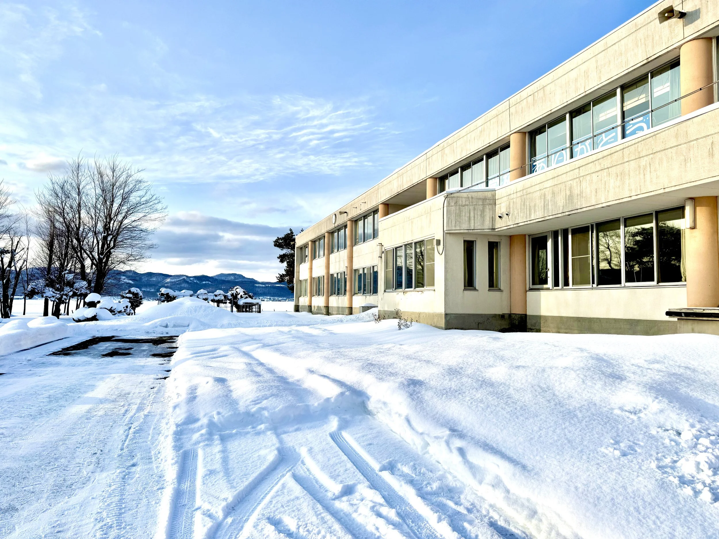 A snow-covered outdoor scene with a modern building featuring large windows, leafless trees, and tracks in the snow, under a partly cloudy sky.