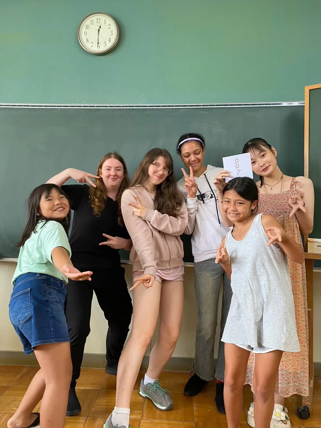 Group of seven diverse young girls posing playfully in front of a blackboard in a classroom with a clock.