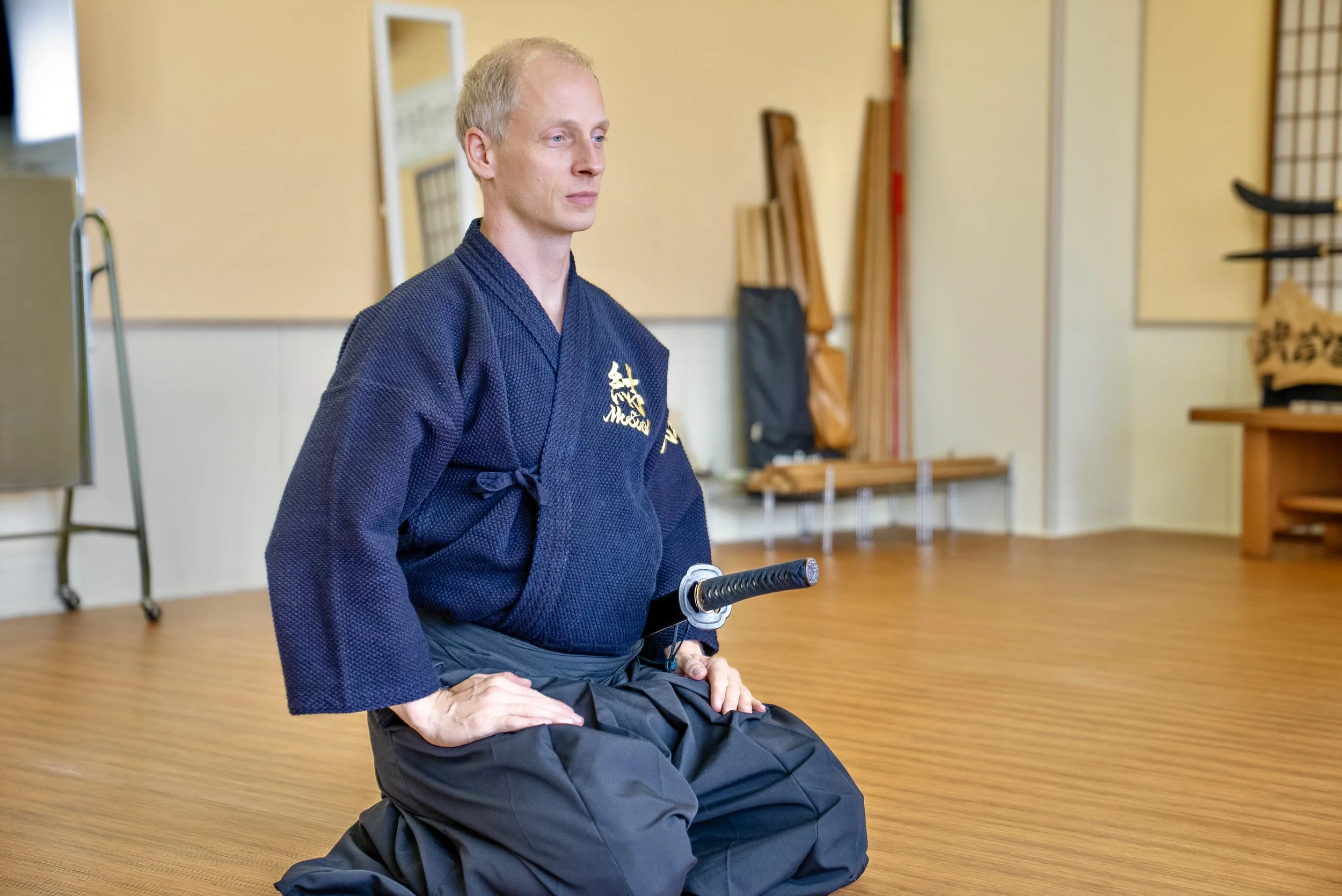 A man in a traditional martial arts uniform kneels in a dojo, holding a katana sword with both hands resting on his lap. The room has wooden flooring, martial arts equipment in the background, and a mirror on the wall.