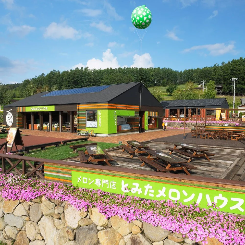 A melon-themed shop with outdoor seating, a stone wall, and a green and white balloon floating above, surrounded by greenery and a blue sky.