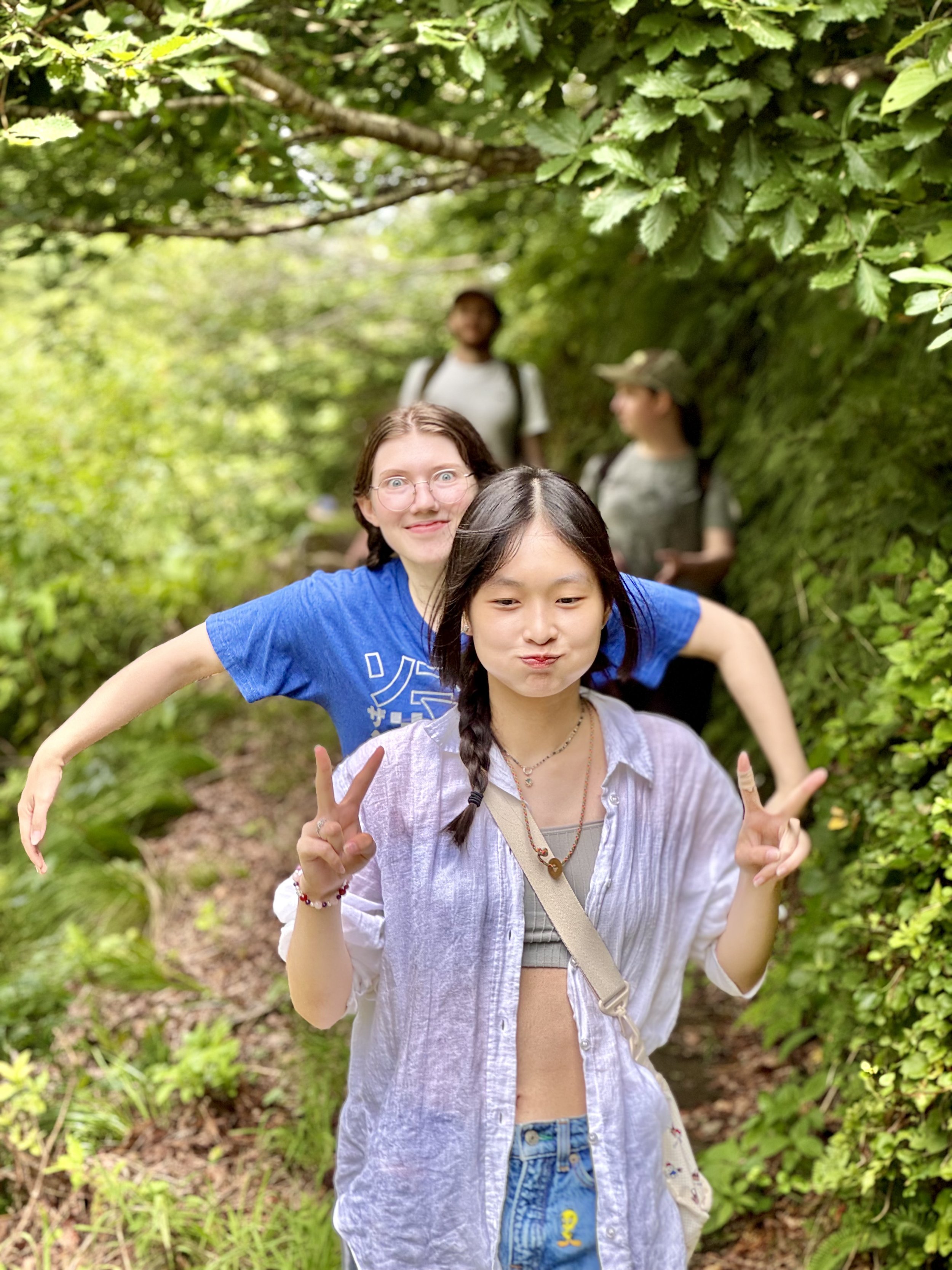 A group of four young women posing playfully on a forest trail surrounded by lush green foliage, with two girls in the front making peace signs and the others smiling and looking into the camera in the background.