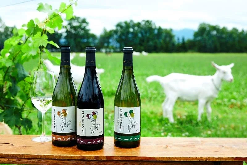 Three bottles of wine with labels, a wine glass filled with white wine, set on a wooden surface outdoors with a green field and white goat in the background