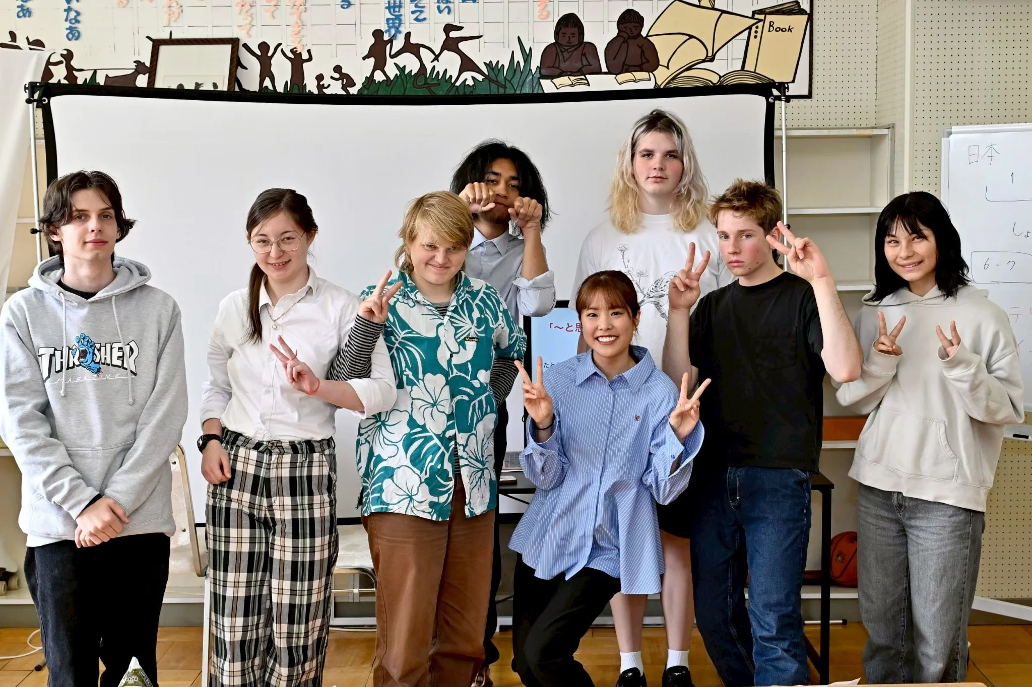 A diverse group of nine students, seven girls and two boys, posing for a photo in what appears to be a classroom. The students are making peace signs or playful gestures. Behind them is a whiteboard and a colorful mural with silhouettes of children playing and reading books.