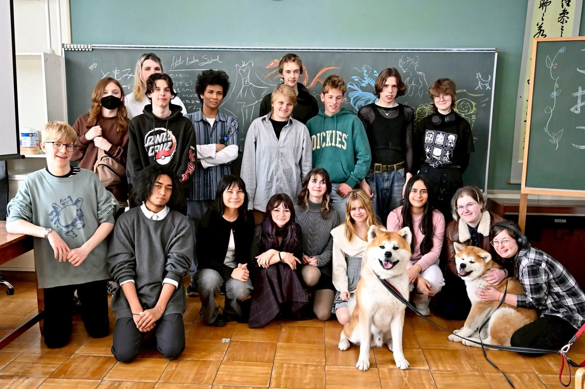 A group of students and two dogs in a classroom, with a chalkboard and drawings in the background.