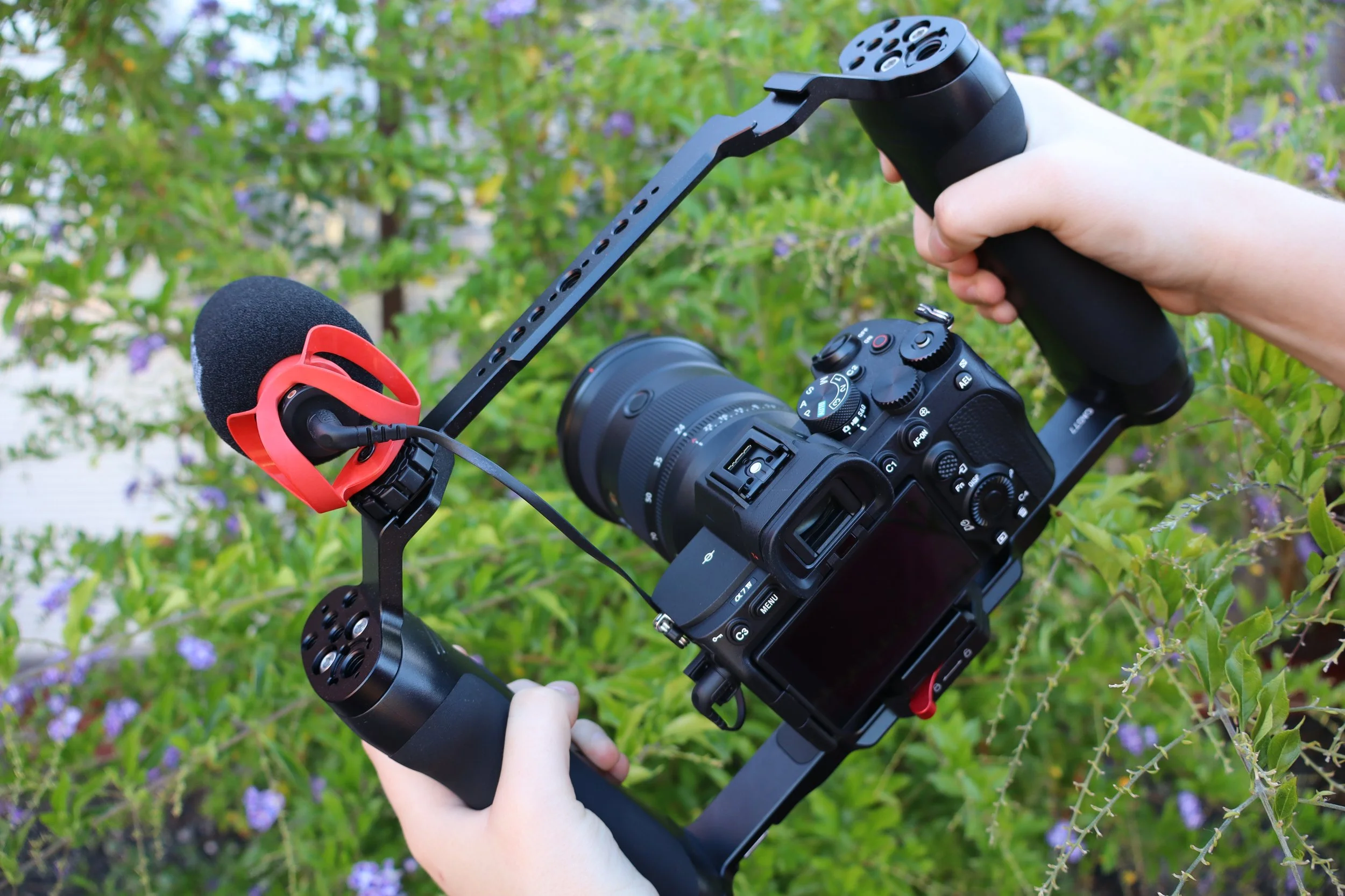A person holding a camera mounted on a stabilizer rig with a microphone attached, outdoors with green foliage and purple flowers in the background.
