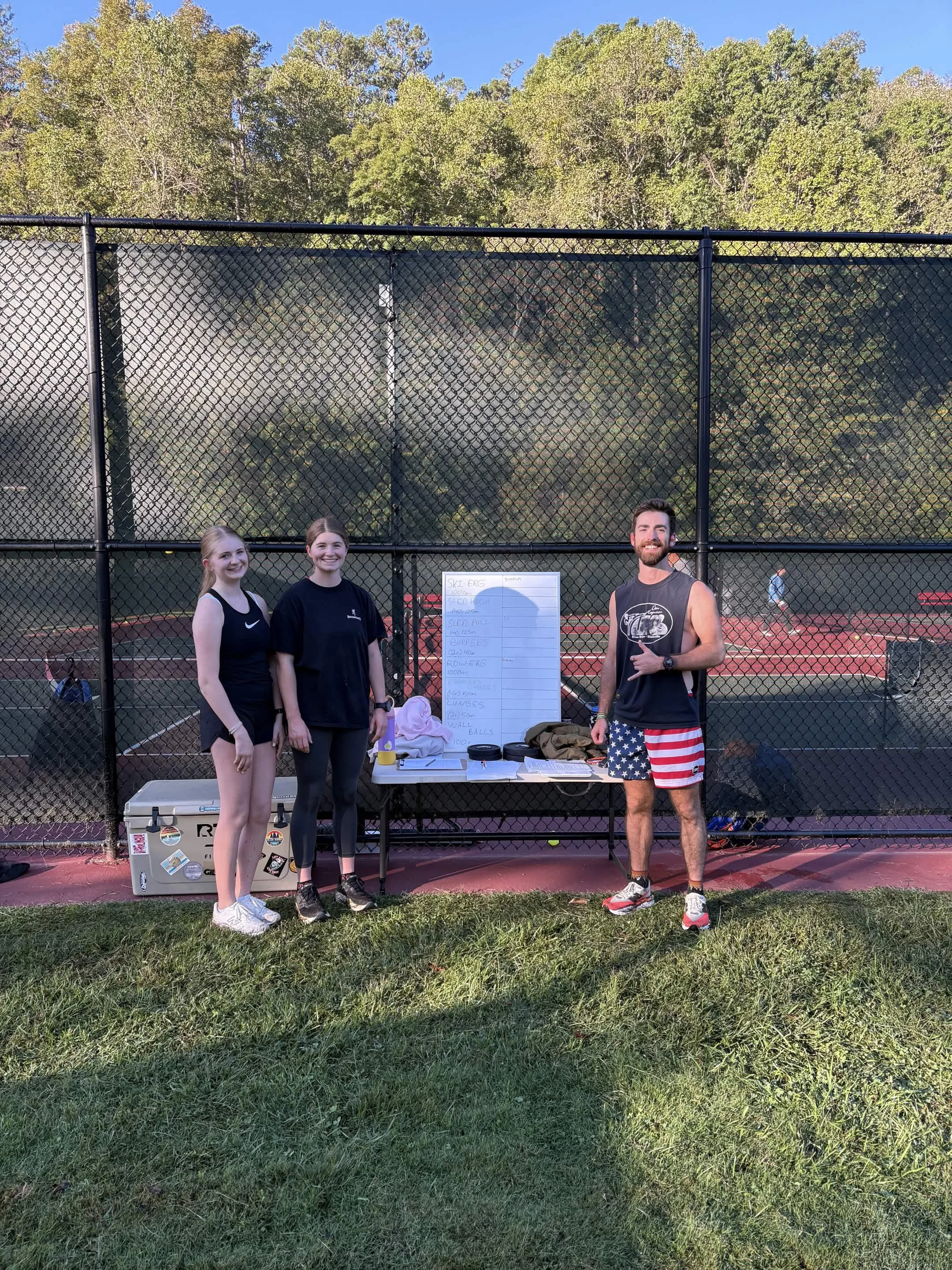 Three people standing on a grassy area in front of a black chain-link fence at a tennis court. Two young women are on the left, and a man on the right. The man is giving a thumbs-up. There is a whiteboard on the table with writing, and a cooler on the ground. The background features trees with green leaves and a clear blue sky.