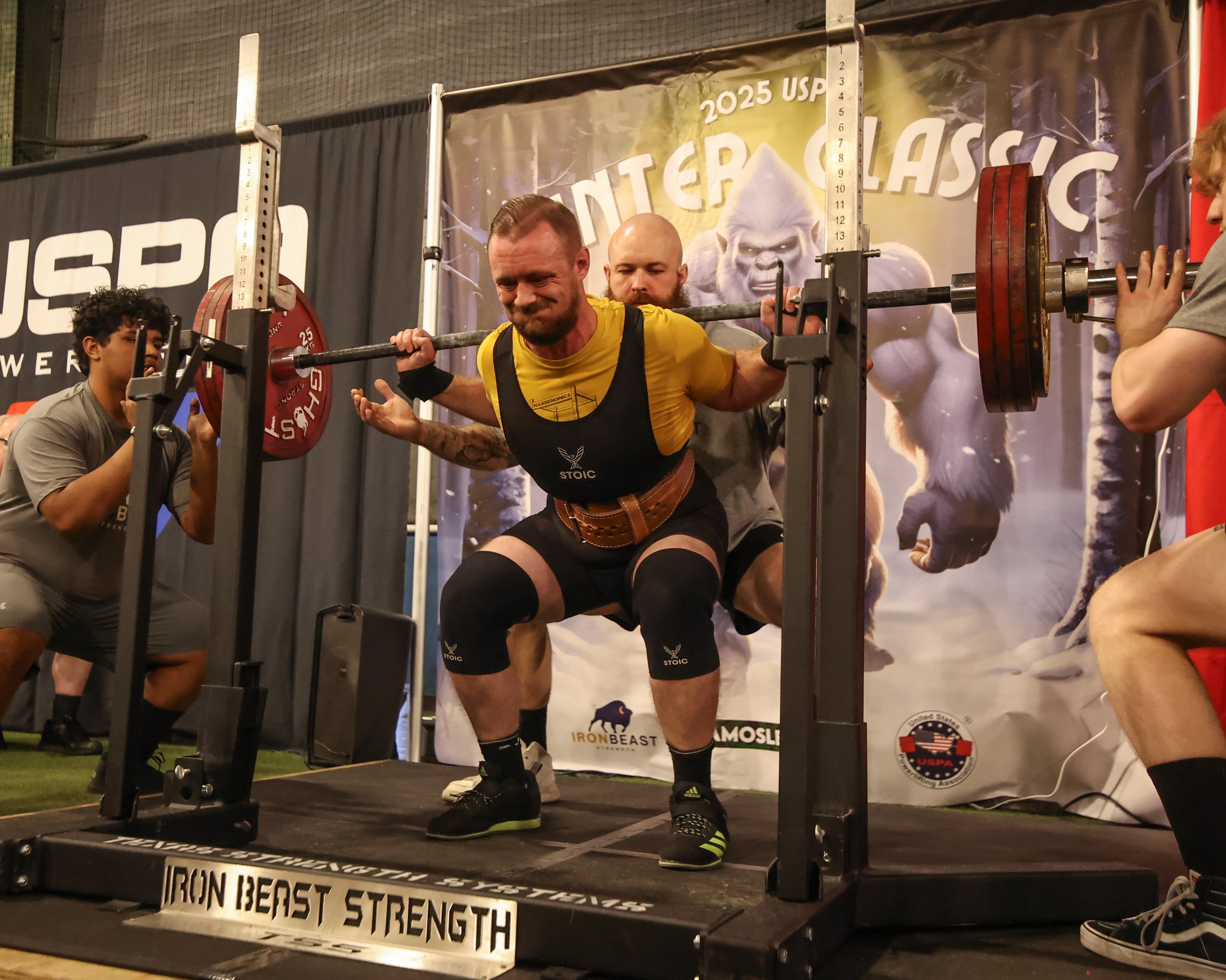 Man in a yellow shirt and black shorts performing a squat at a powerlifting competition, with spotters on each side and supporting equipment including a barbell with red weight plates. The background has a banner with a gorilla image and the text '2025 USPA Winter Classic'.