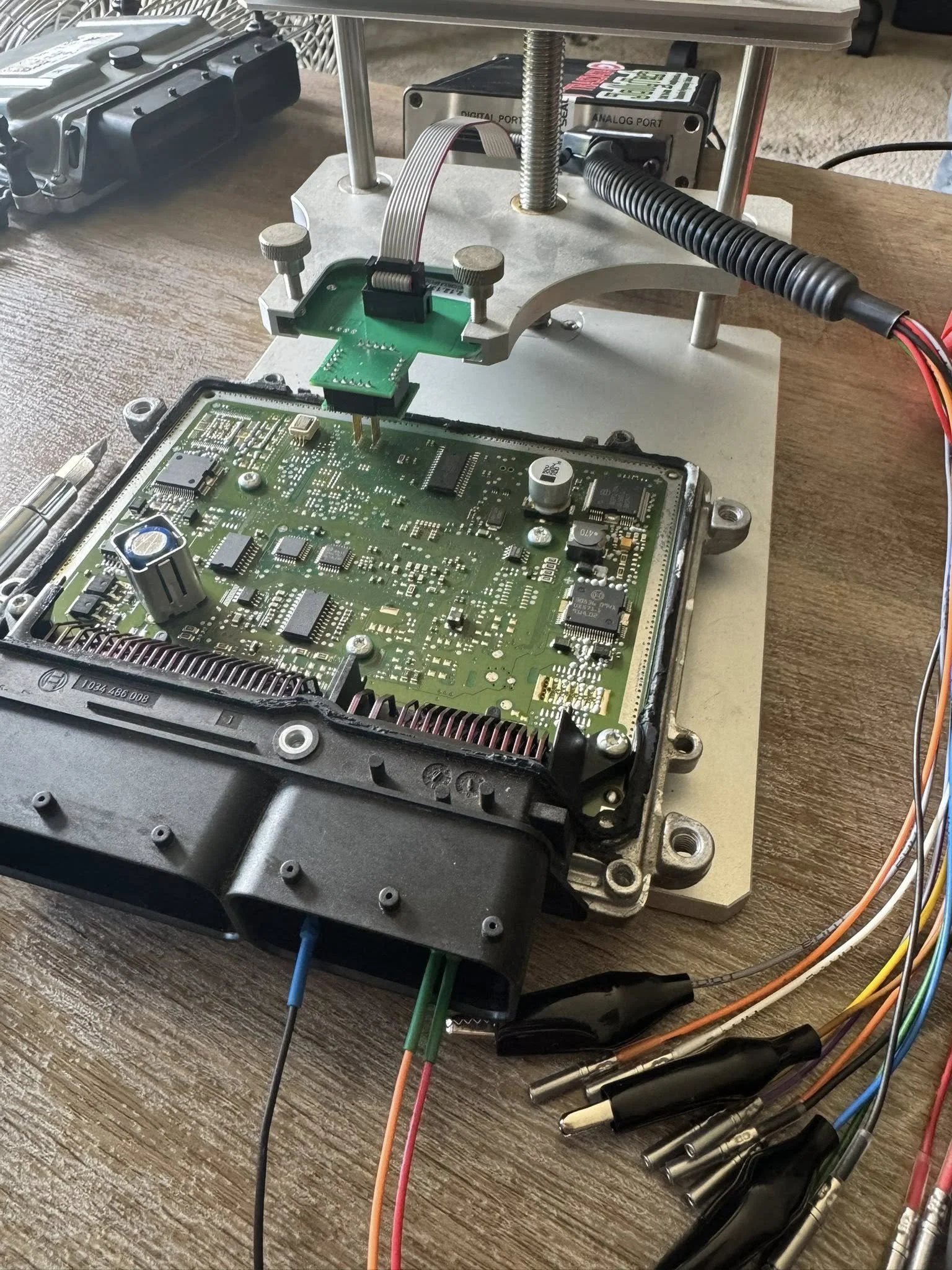 Close-up of a circuit board with various electronic components, connected to testing equipment with wires and clips, on a wooden table.