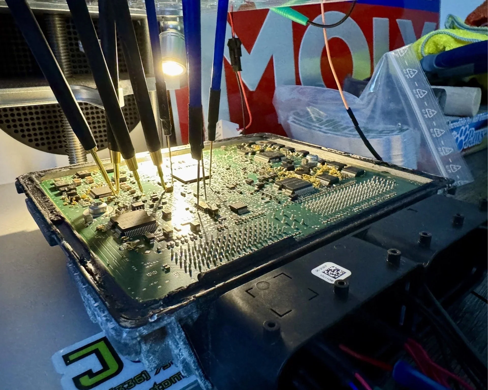 Close-up of an electronic circuit board on a workbench, with multiple soldering probes and a bright light illuminating the board. There are various electronic components and small pins on the circuit board.
