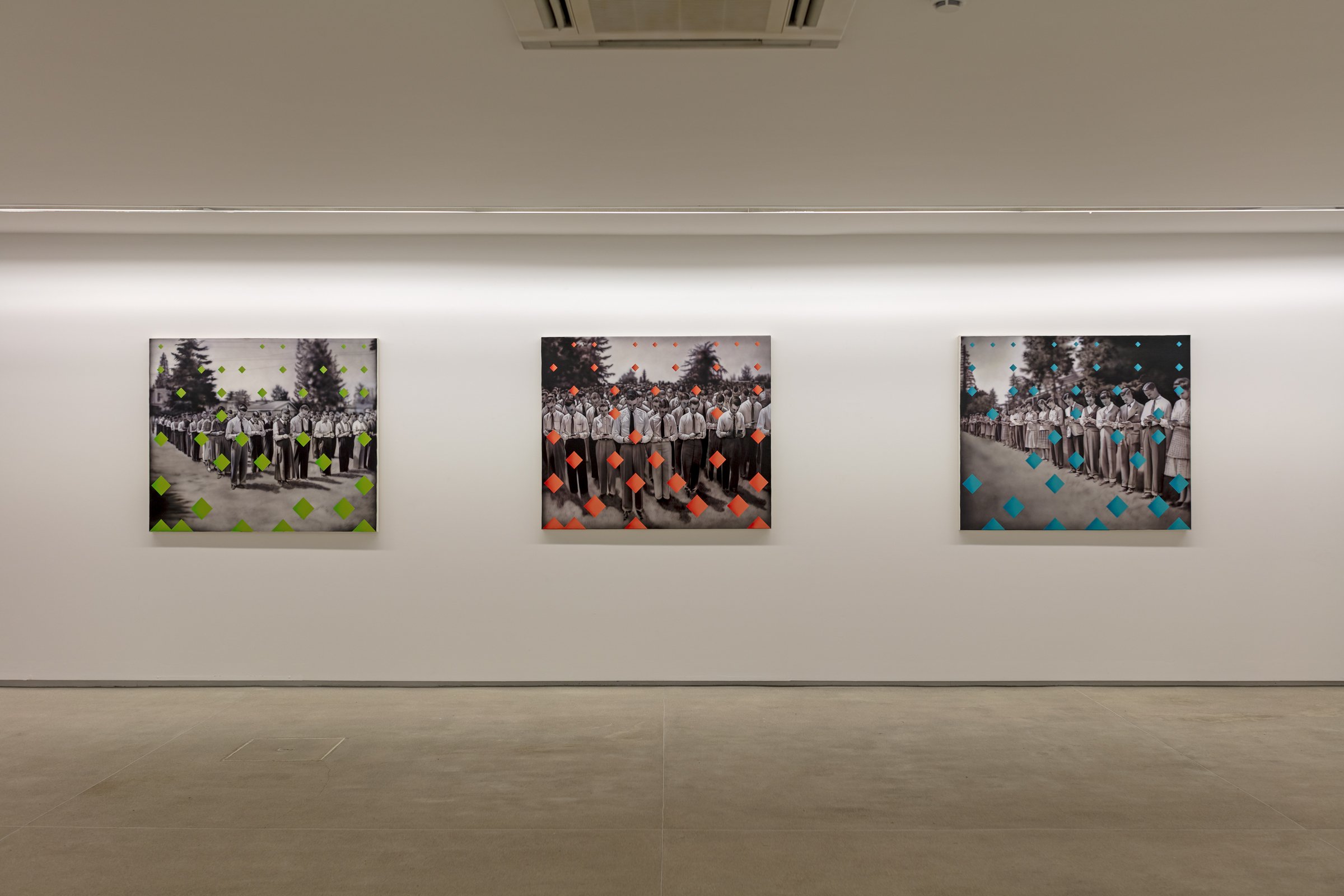 Tres cuadros en una galería, cada uno presenta una fotografía en blanco y negro de un grupo de personas paradas en fila, con puntos de colores superpuestos: en el primero puntos verdes, en el central puntos naranjas, en el tercero puntos azules.