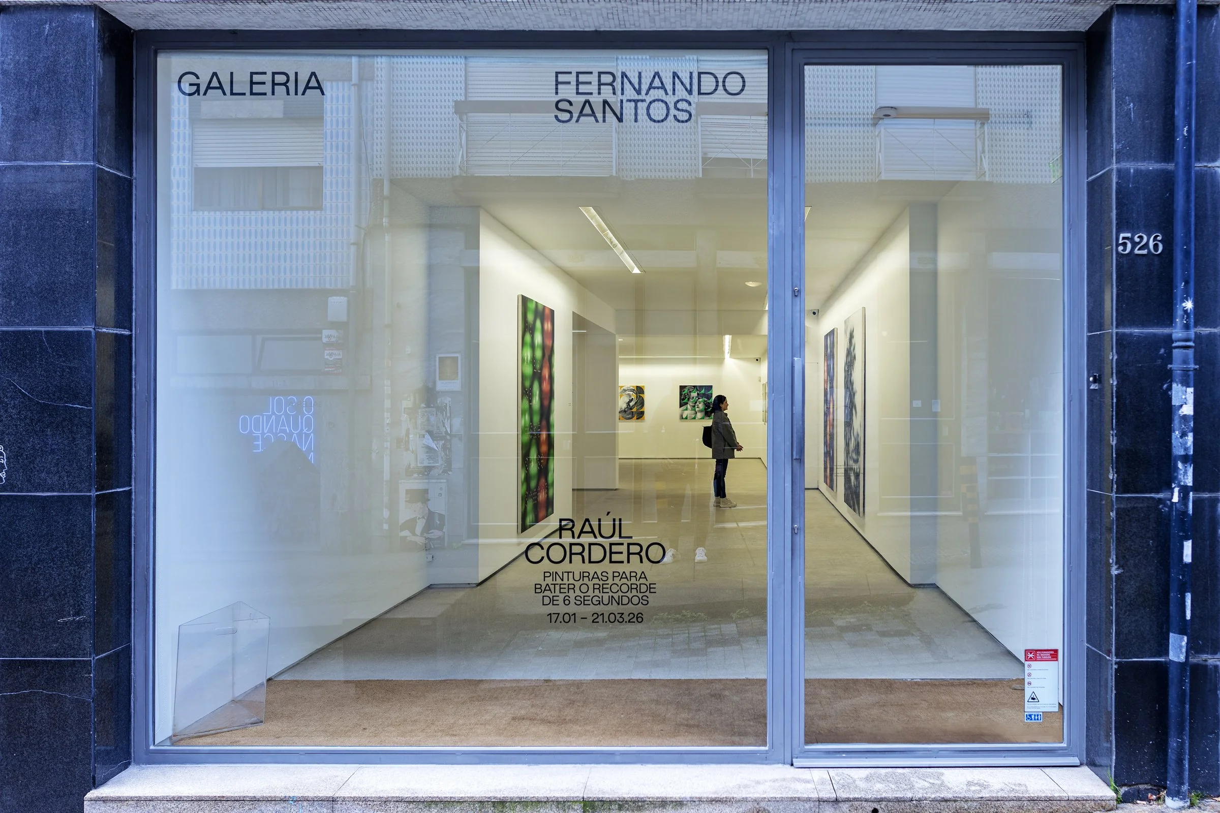 Vista de una galería de arte con una ventana de cristal en la entrada, mostrando obras de arte en las paredes y una persona observando las pinturas en el interior.