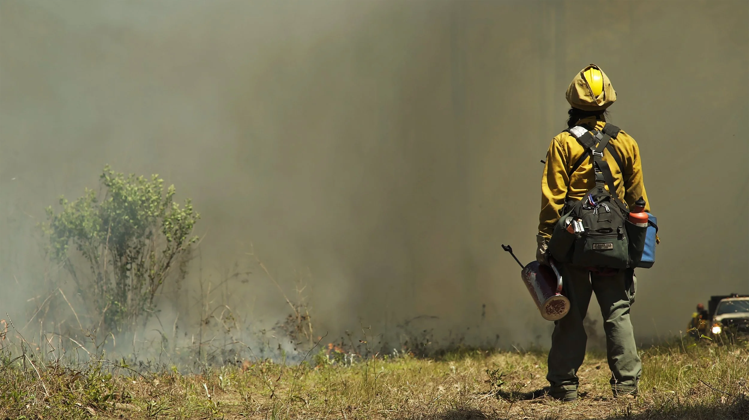 A firefighter on a grassy field, with a backpack and protective gear, looks at a smoky wildfire in the distance.