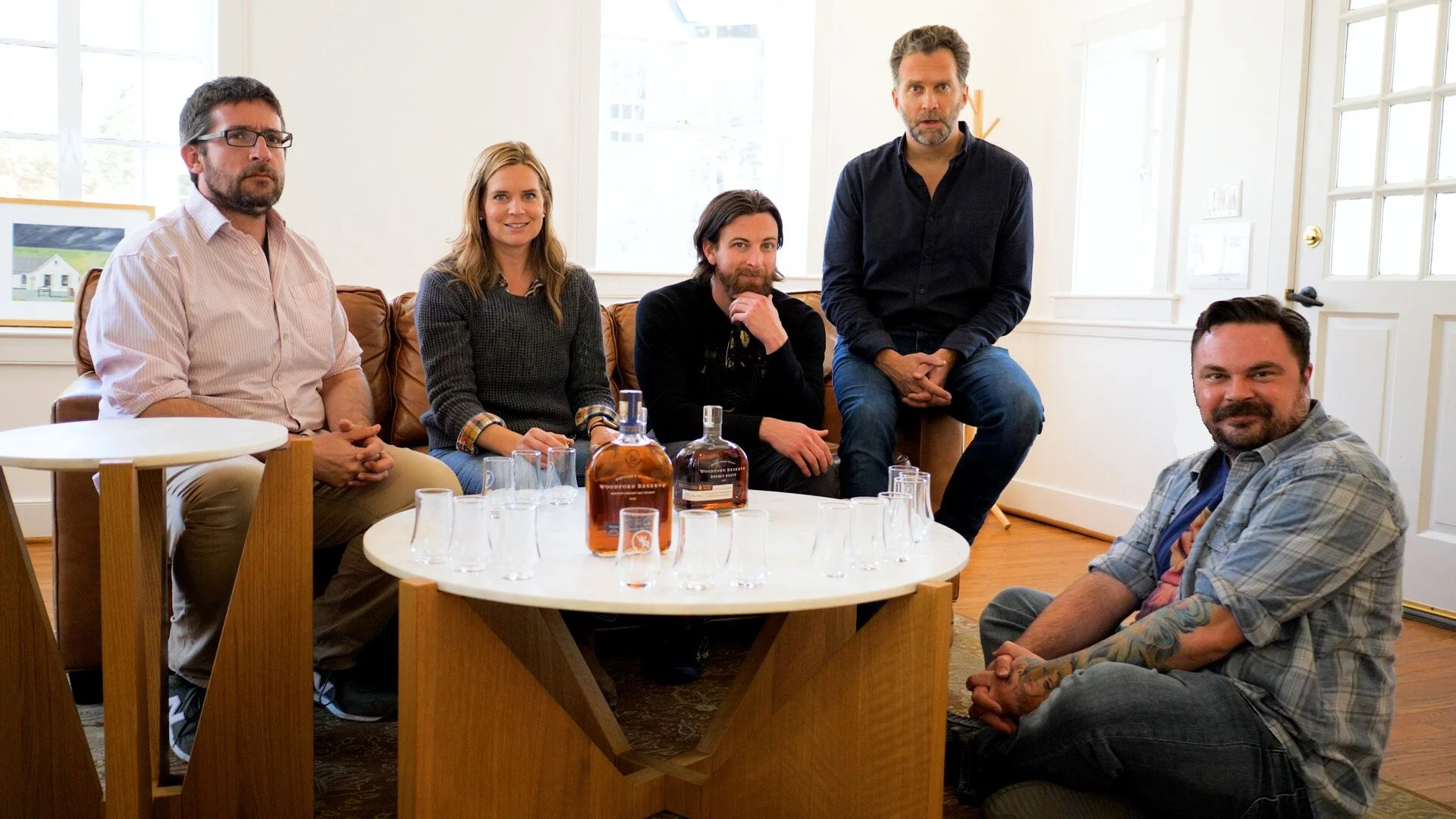 Six people, three men and three women, gathered around a table with whisky bottles and glasses, in a bright room with windows and artwork, posing for a photo.