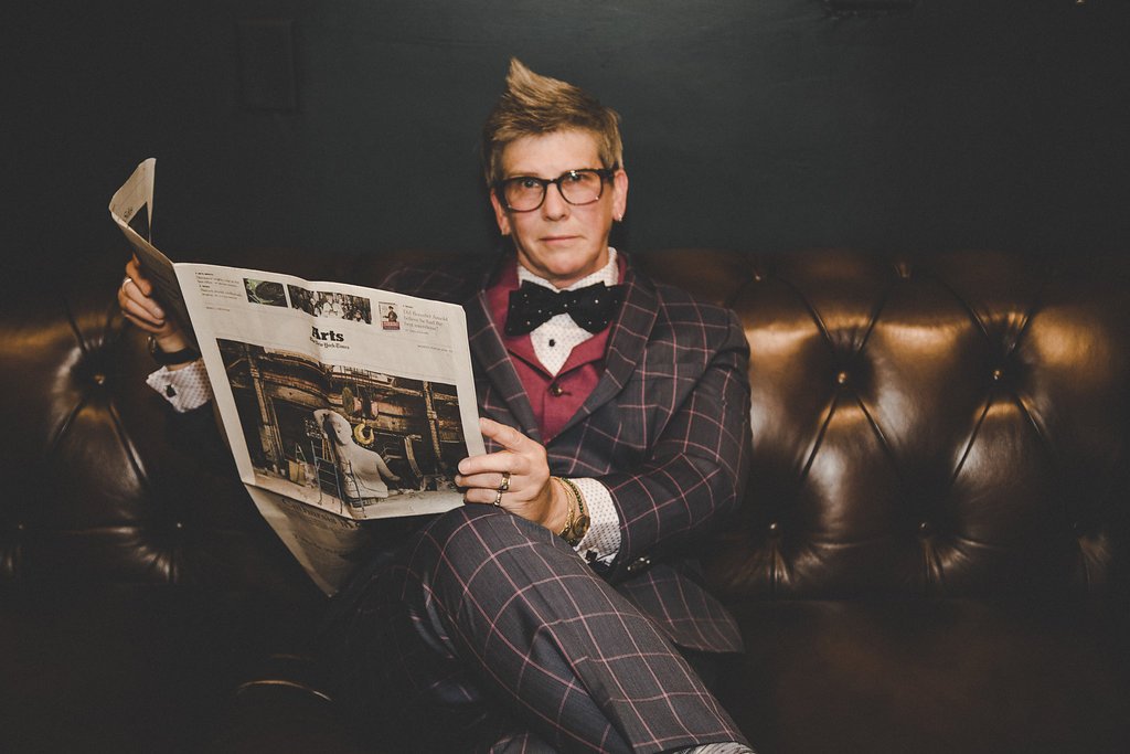 A man with glasses and a patterned suit sitting on a leather couch, reading a newspaper.