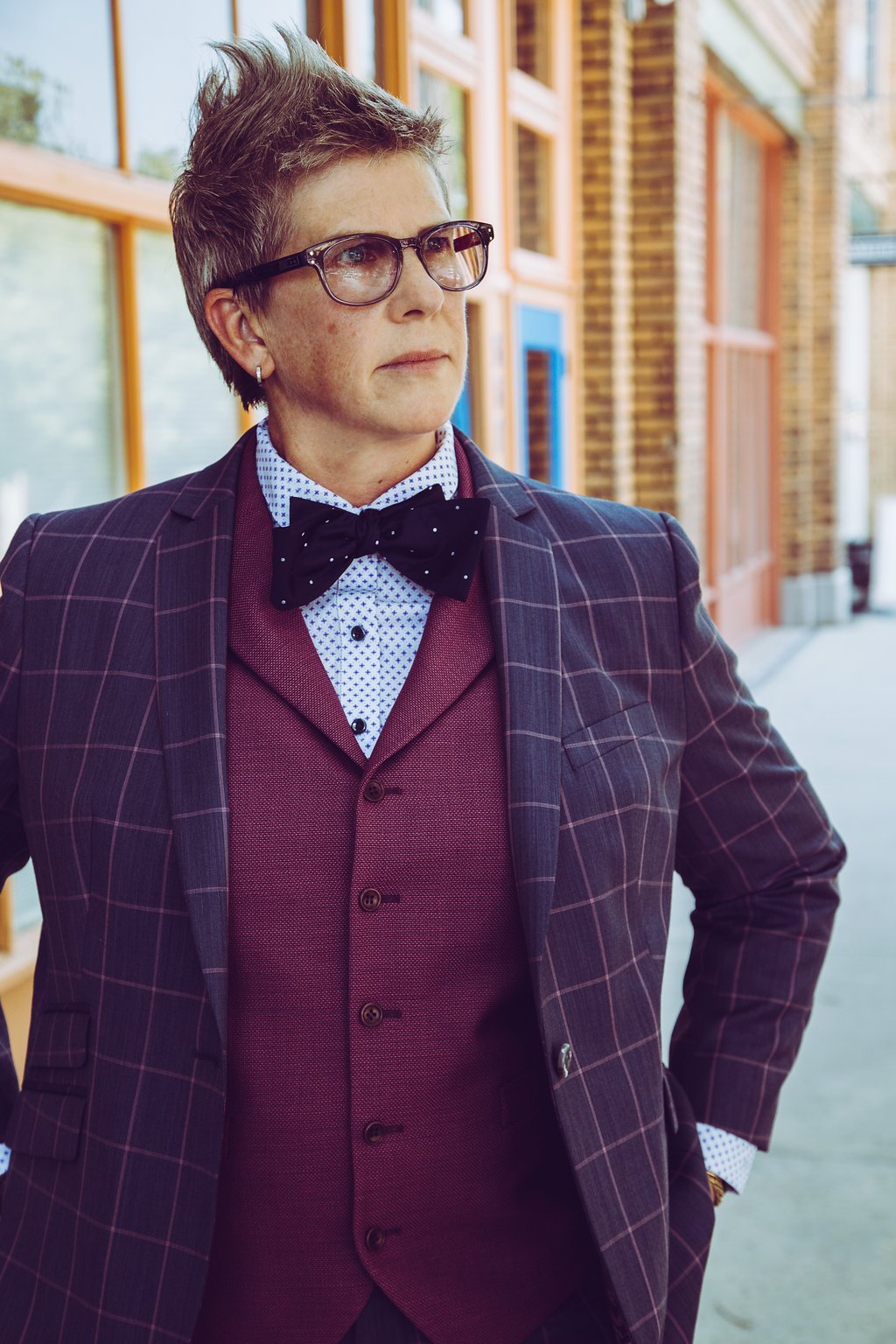 A person with short, spiky hair, glasses, and earrings wearing a purple pinstripe suit jacket, a purple vest, a polka dot shirt, and a black bow tie with white dots, standing outdoors near a brick building with large windows.