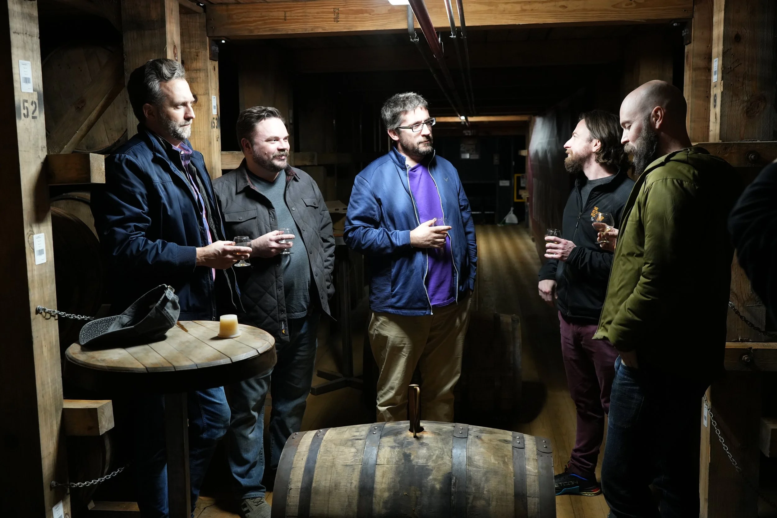 Six men standing in a rick house, holding glasses of bourbon, engaged in conversation. One man is talking to the others while the rest listen.  The men are surrounded by bourbon barrels and a small round wooden table with a hat and a candle.