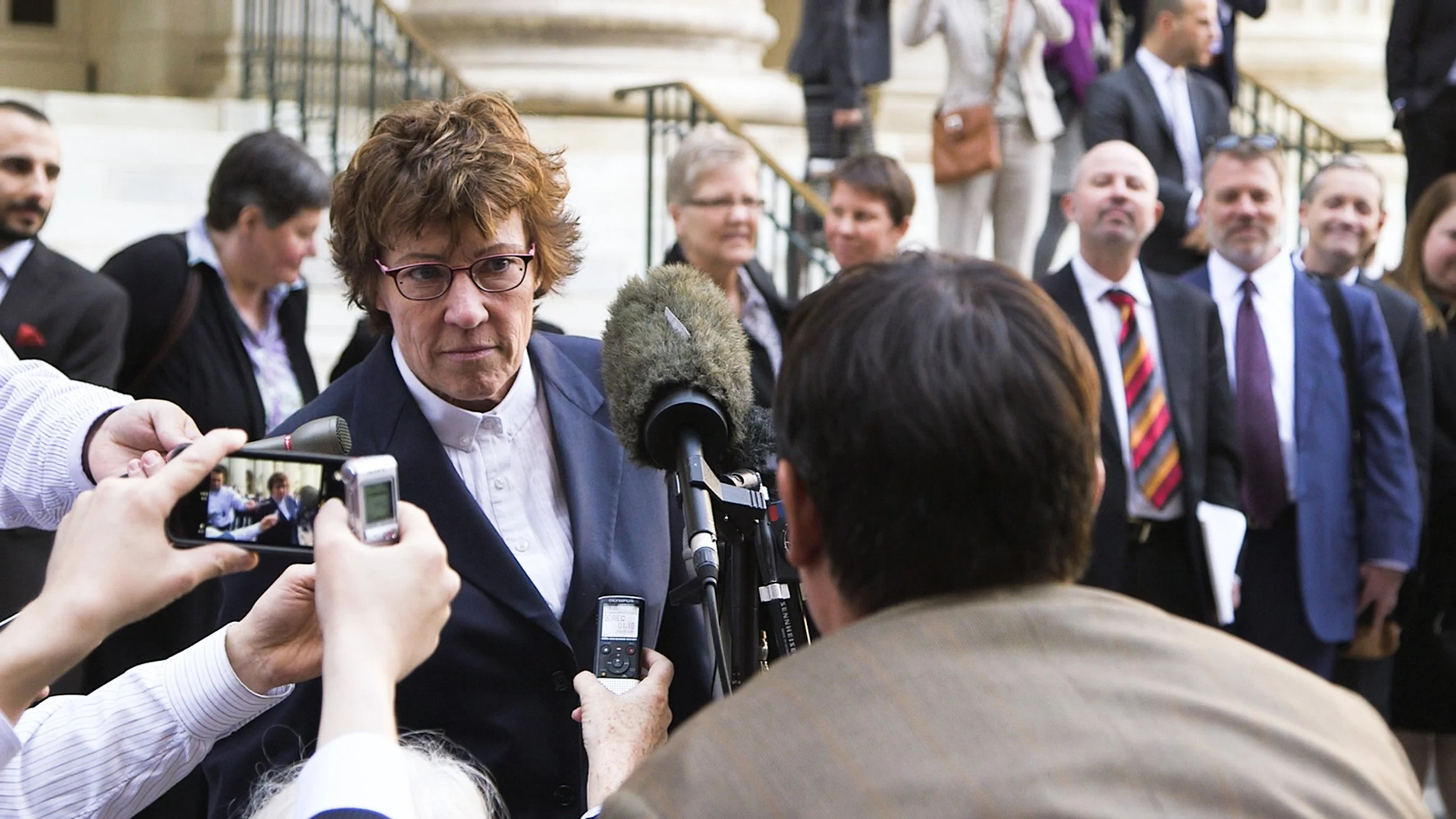 A woman with short brown hair, glasses, and a navy blazer being interviewed outside, with multiple people standing in the background.