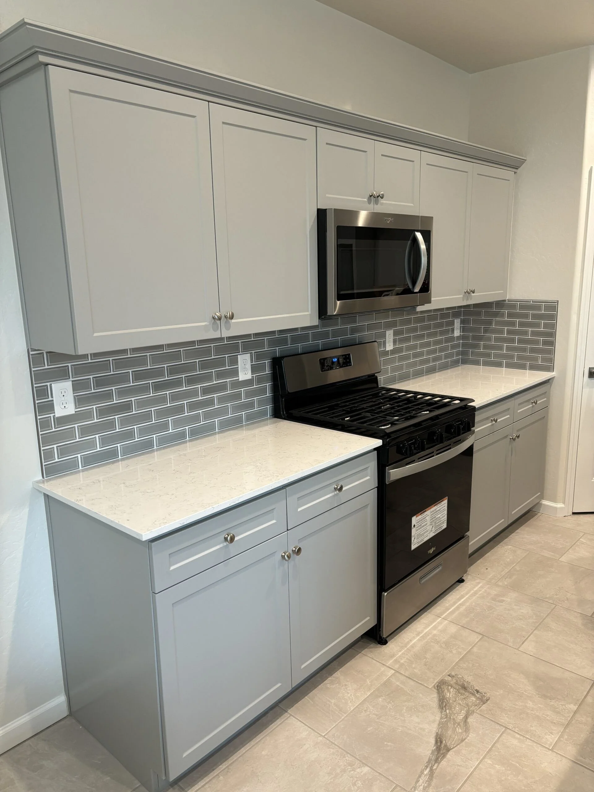 Kitchen with light gray cabinets, gray subway tile backsplash, white speckled countertop, stainless steel microwave, and stove.