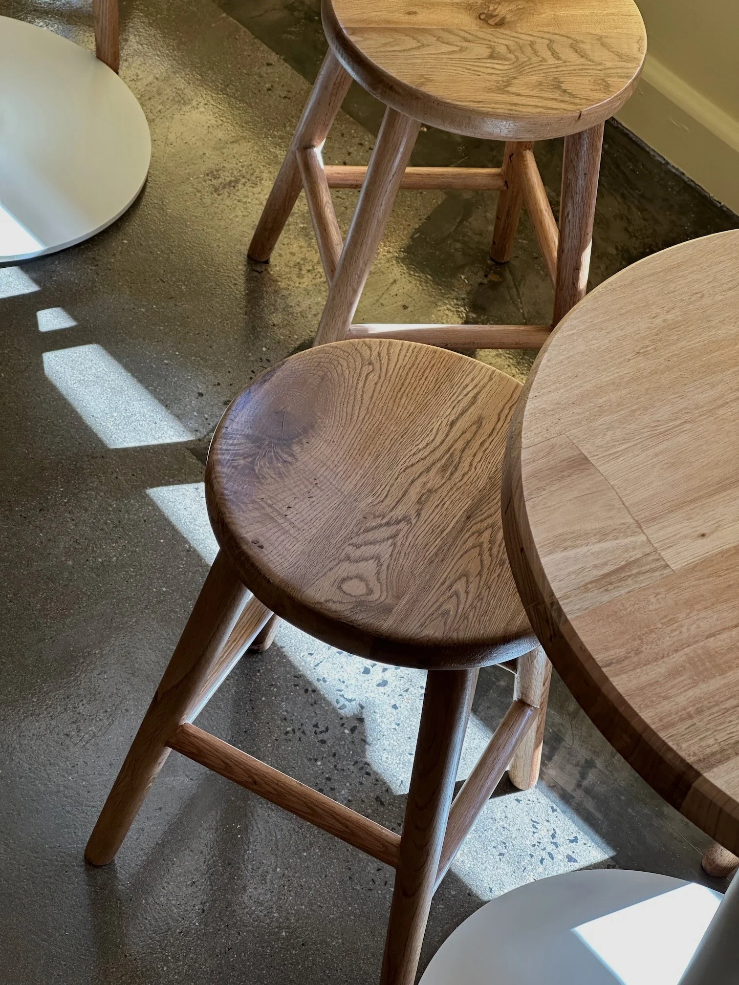 Wooden chair next to a round wooden table, with sunlight casting shadows on a concrete floor.
