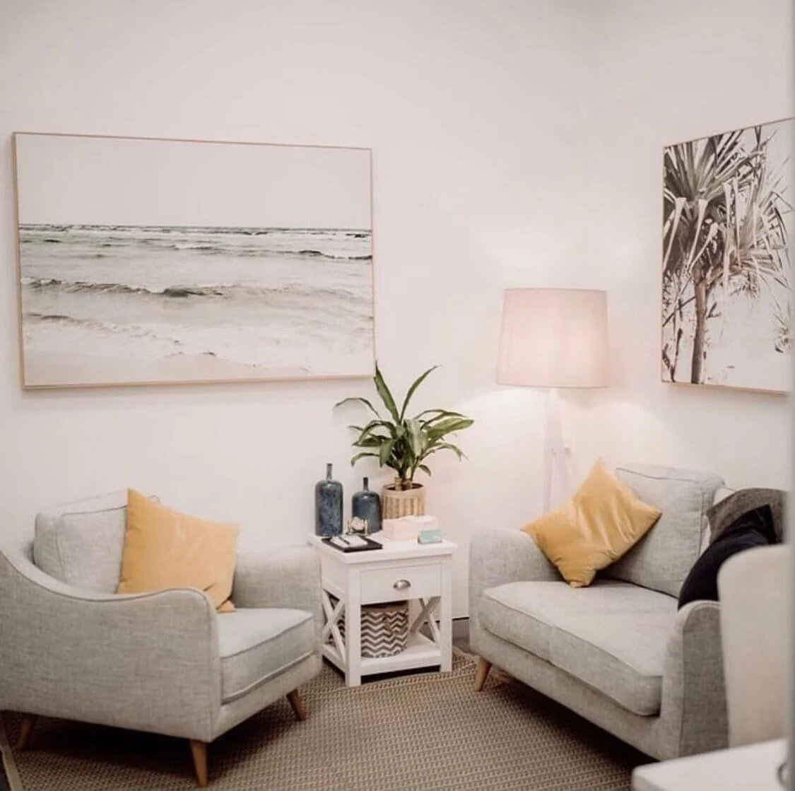 Bright coastal living room with gray armchair and sofa, mustard yellow pillows, white side table, potted plant, floor lamp, and ocean-themed wall art