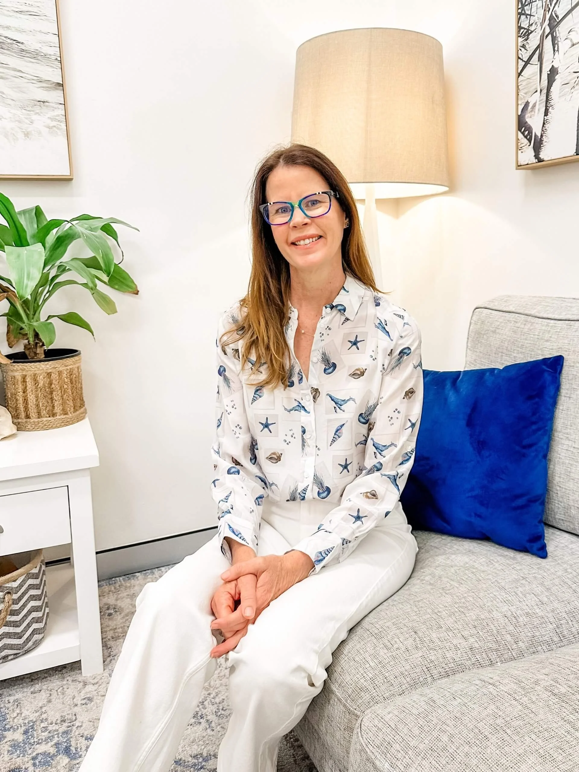 Smiling woman wearing blue glasses and a seashell-print blouse seated on a gray sofa beside a lamp and potted plant