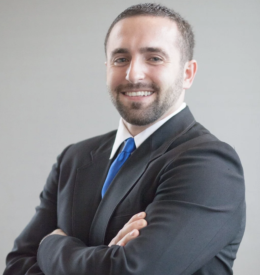 A man wearing a black suit, white shirt, and blue tie, smiling with arms crossed, standing against a plain light gray background.