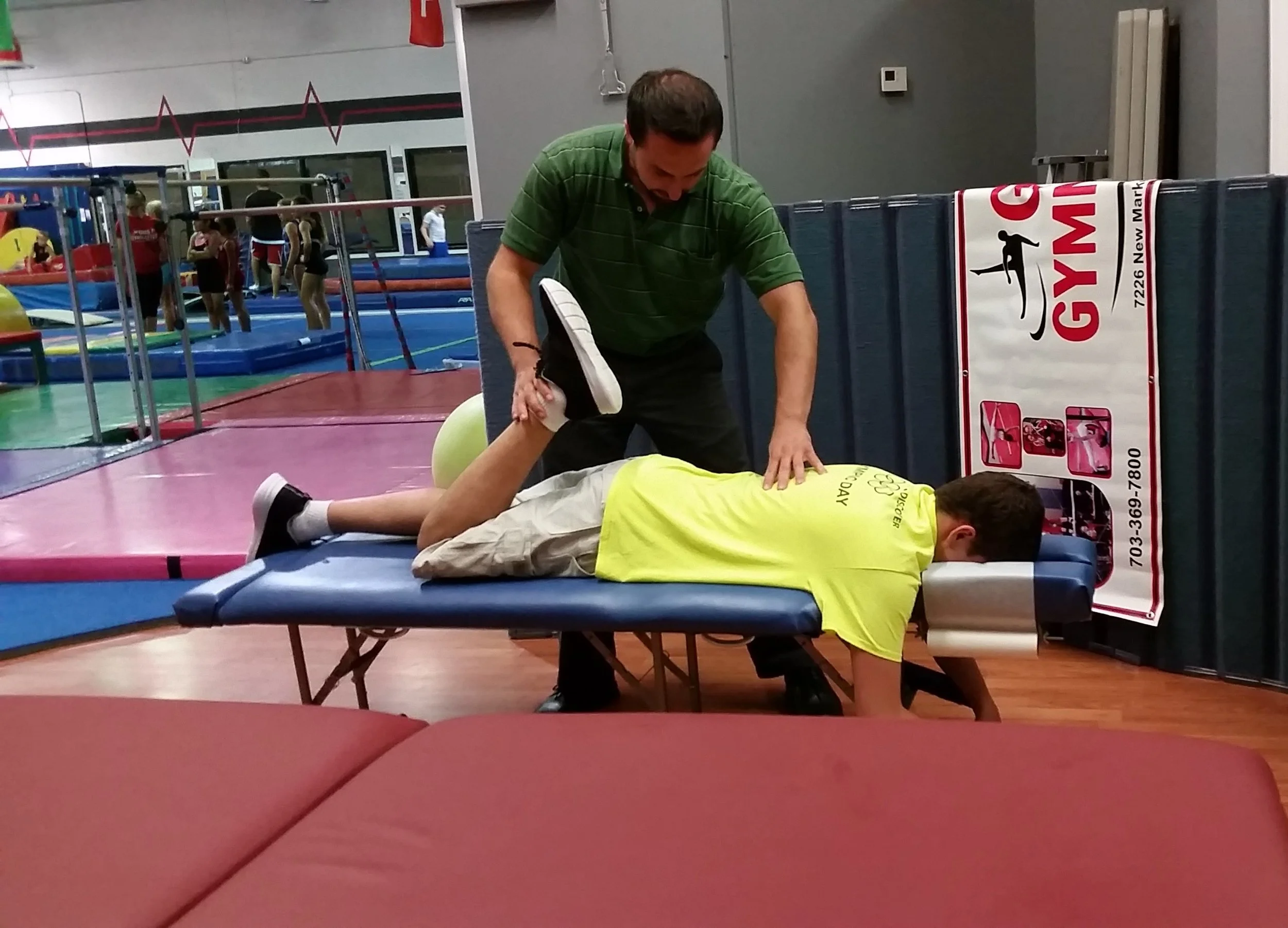 A person lying face down on a massage table while another person helps stretch their leg at a gymnastics gym.