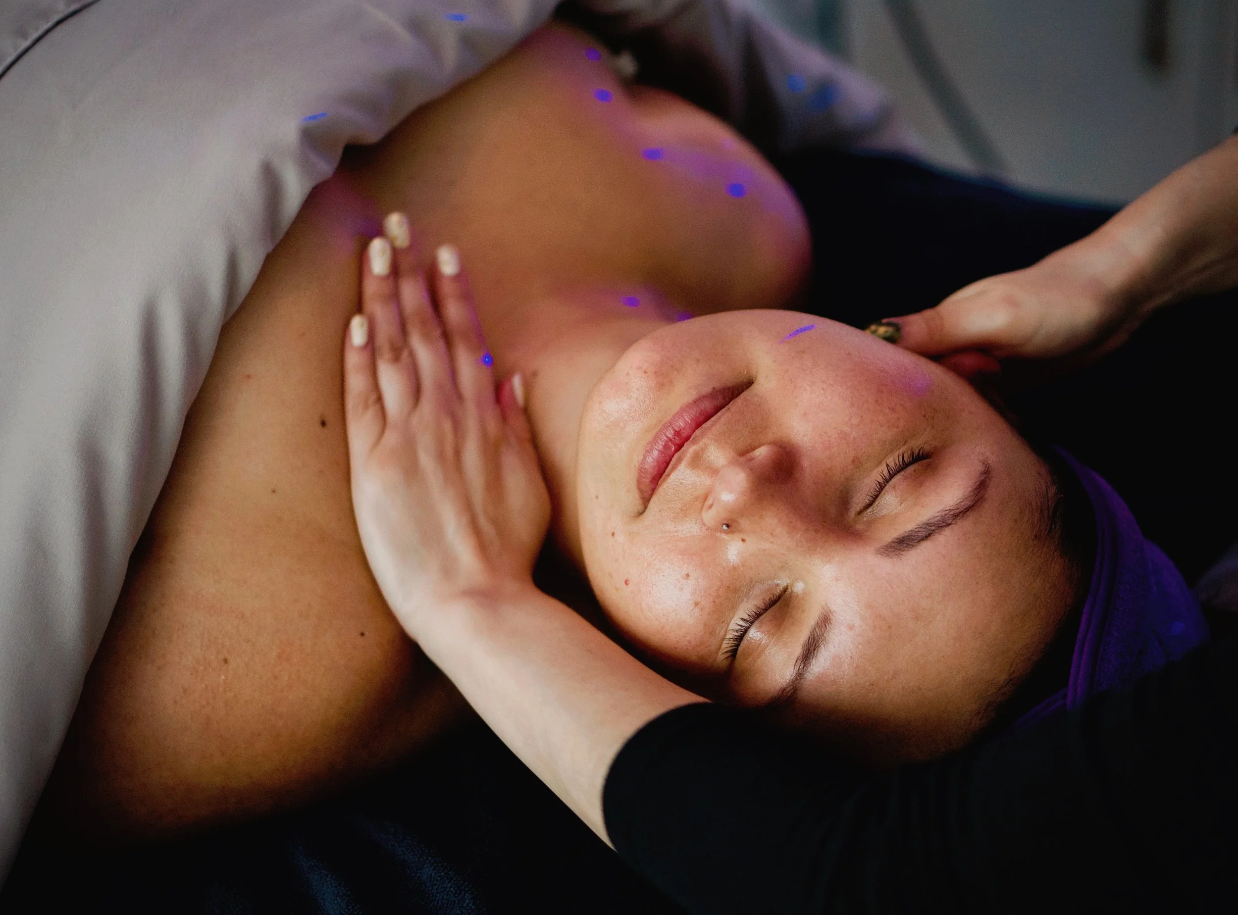 A woman receiving a massage, lying on her back with eyes closed, head resting on a pillow, with purple lighting on her face and shoulder.
