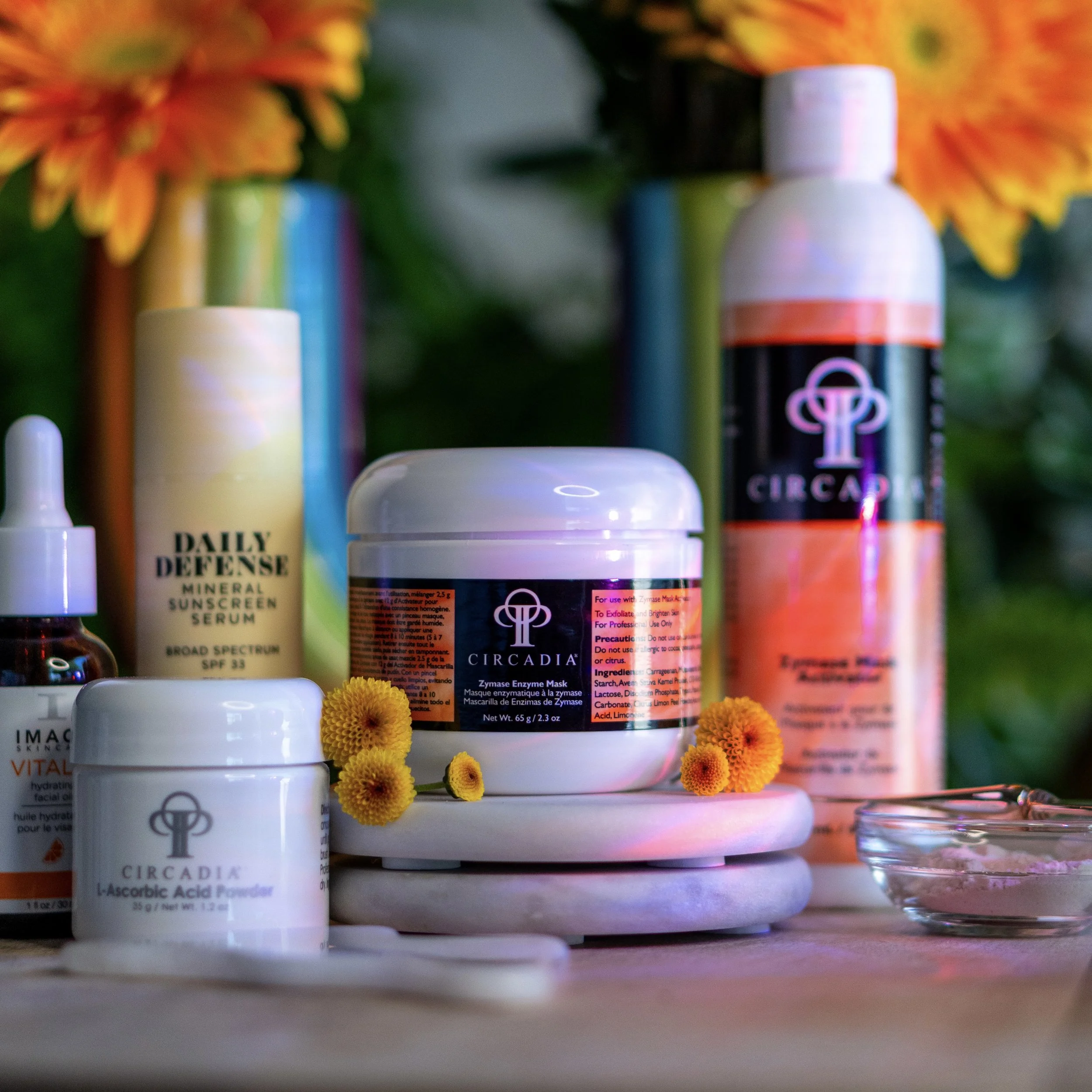 Skincare products on a white surface with yellow flowers and colorful vases in the background, including Circadia enzyme mask and L-ascorbic acid powder.