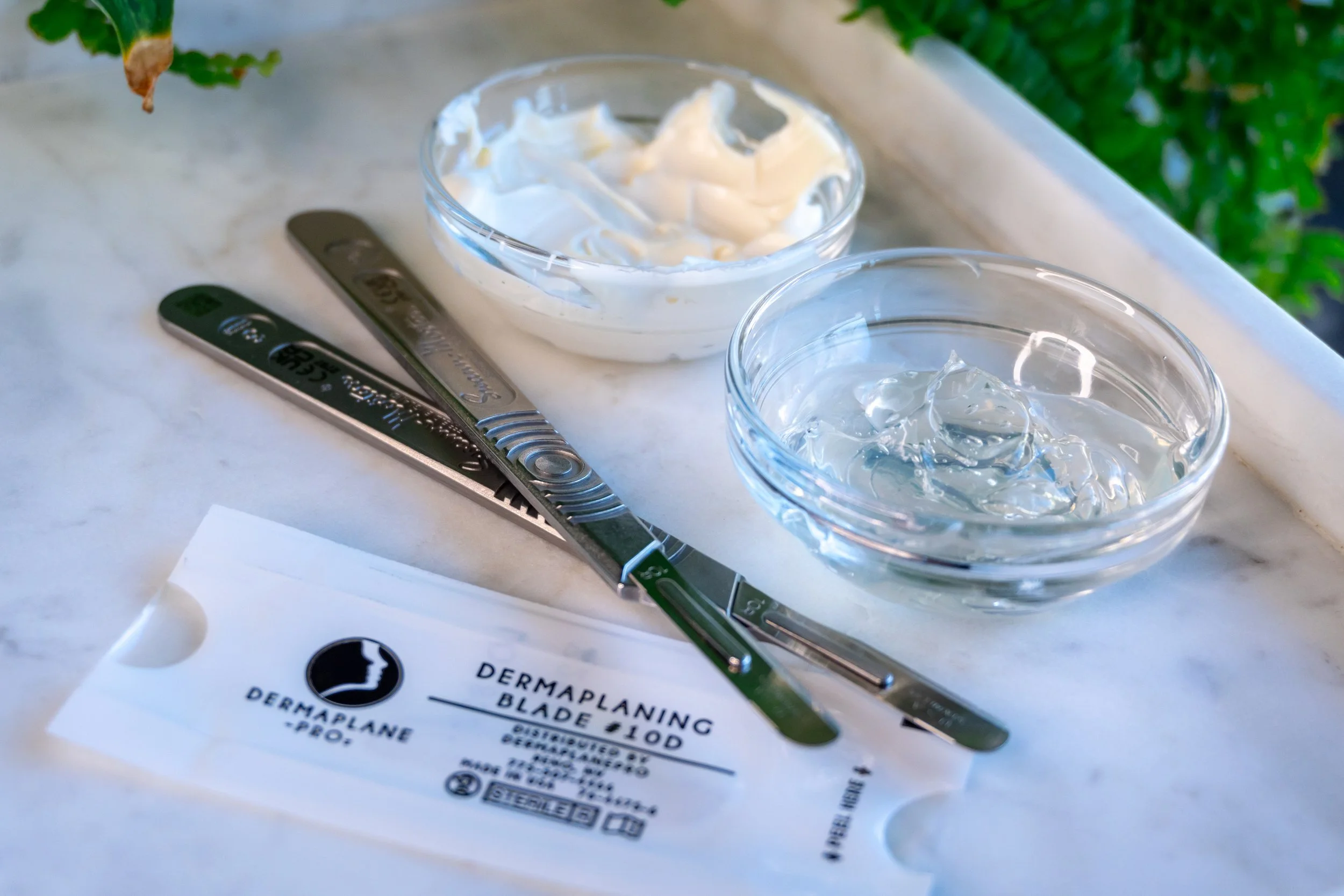 skin care tools including a dermaplaning blade, a metal spatula, and two glass bowls with creams, placed on a white surface.