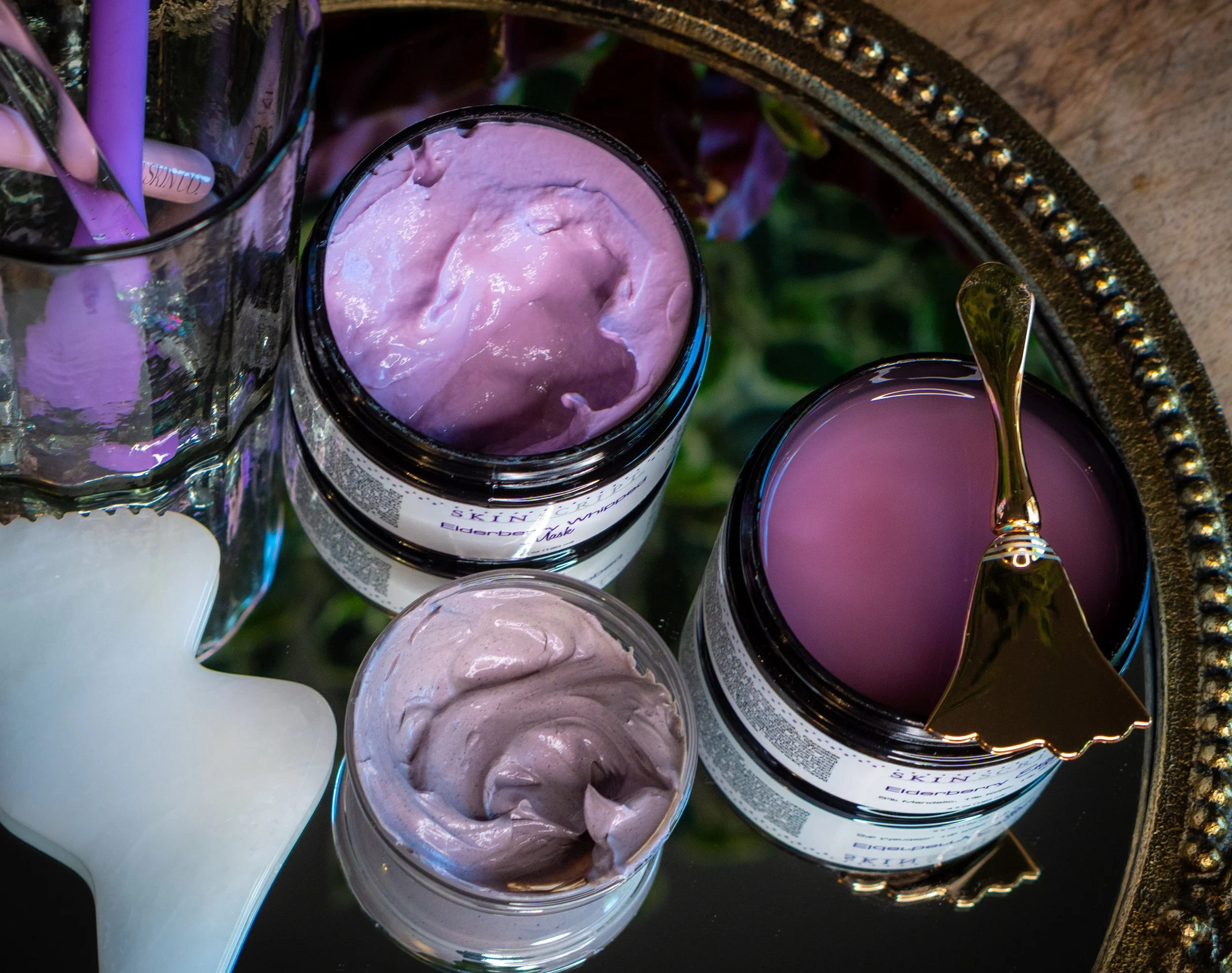 Beauty products, including pink and purple creams in jars, a white candle, and a gold spoon, placed on a mirrored tray.