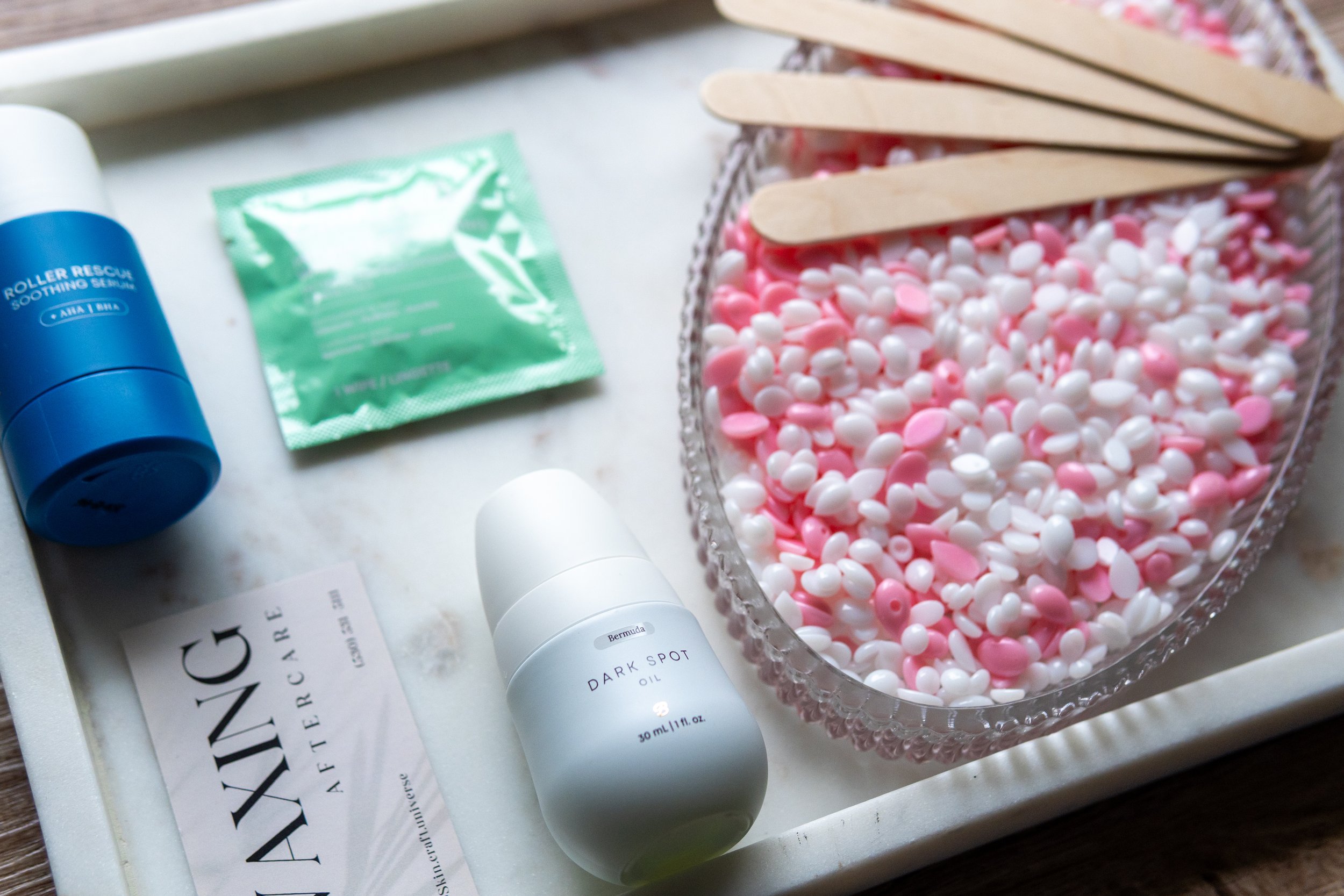 A tray with various skincare and wellness items, including a blue roller rescue soothing balm, a green packet, a white bottle labeled 'Dark Spot Oil,' a card with partially visible text, a bowl filled with pink and white candy sprinkles, and several wooden sticks.