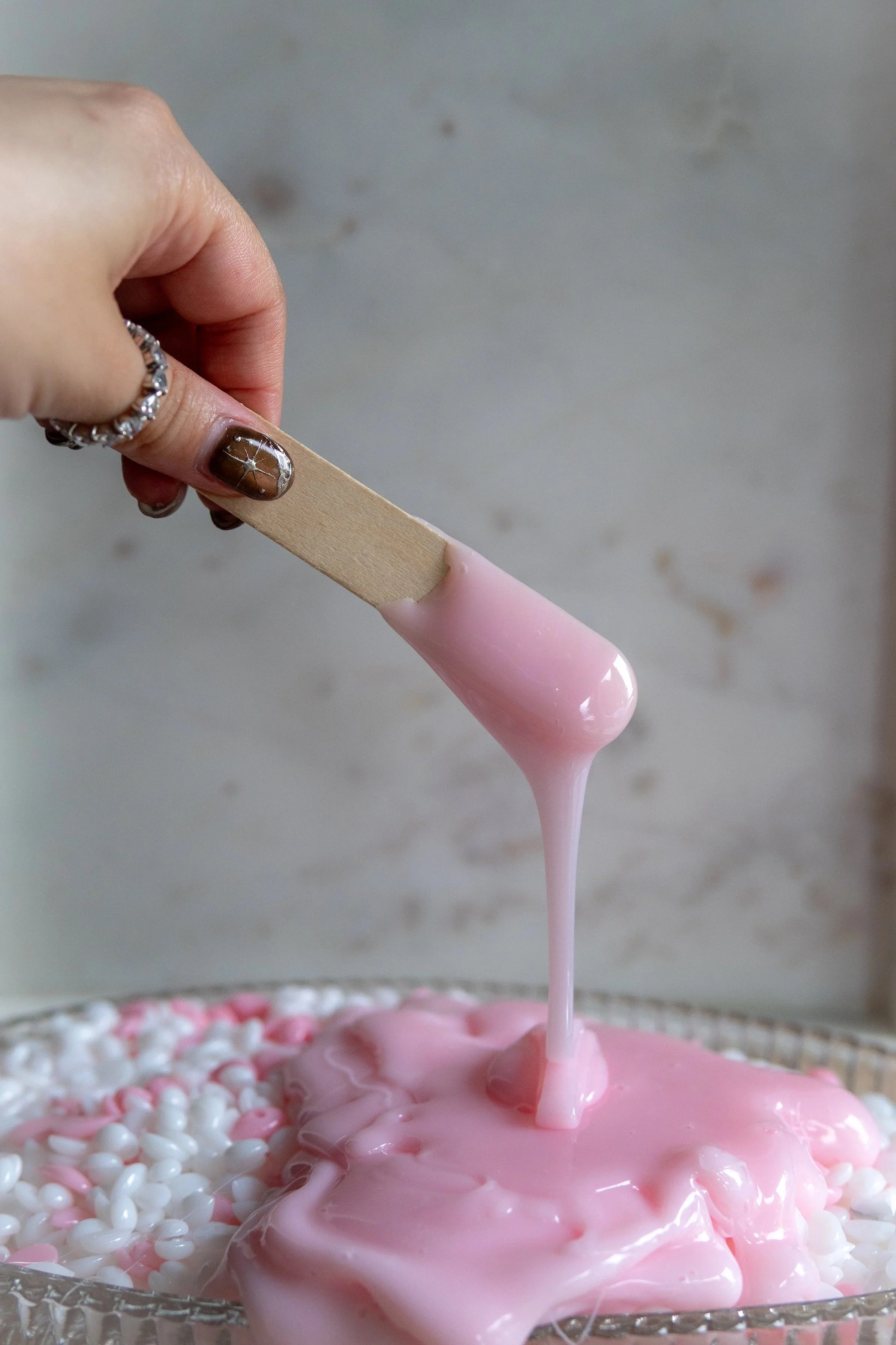 Person holding a wooden stick with pink slime dripping from it over a container of white and pink beads.