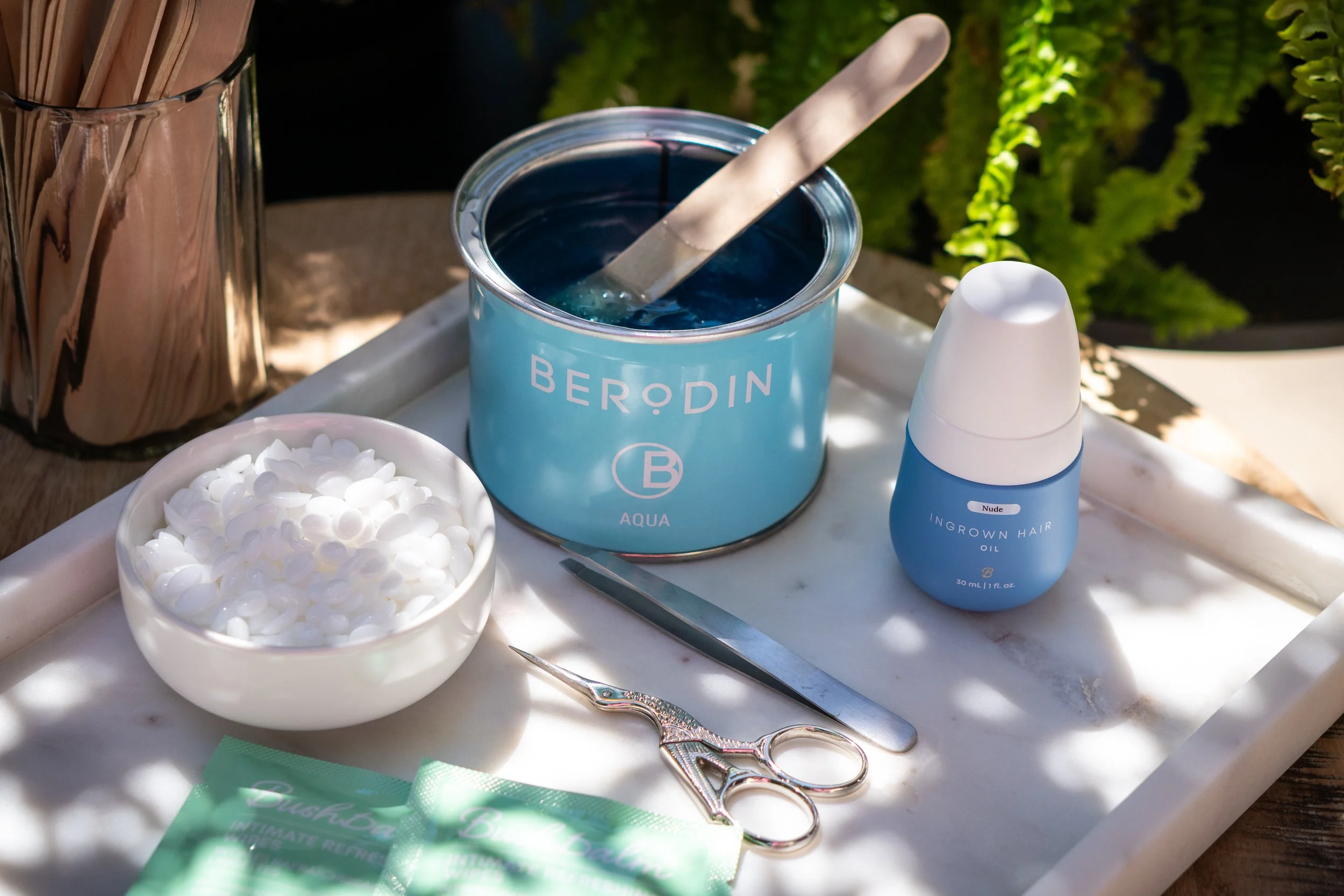 A tray of skincare and hair removal tools, including a container of blue wax, a bowl of white waxing strips, a pair of small scissors, a spatula, and a bottle of ingrown hair oil, set outdoors on a marble surface with plant greenery in the background.