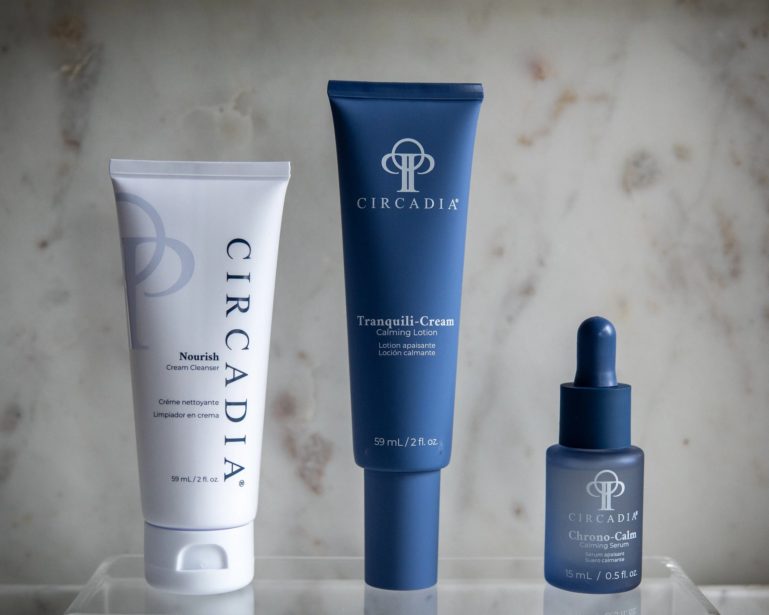 Three skincare products: a white tube labeled Nourish Cream Cleanser, a blue tube labeled Circadia Tranquili-Cream Calming Lotion, and a small blue bottle labeled Circadia Chrono-Calm Calming Serum, placed on a clear surface against a beige marble background.
