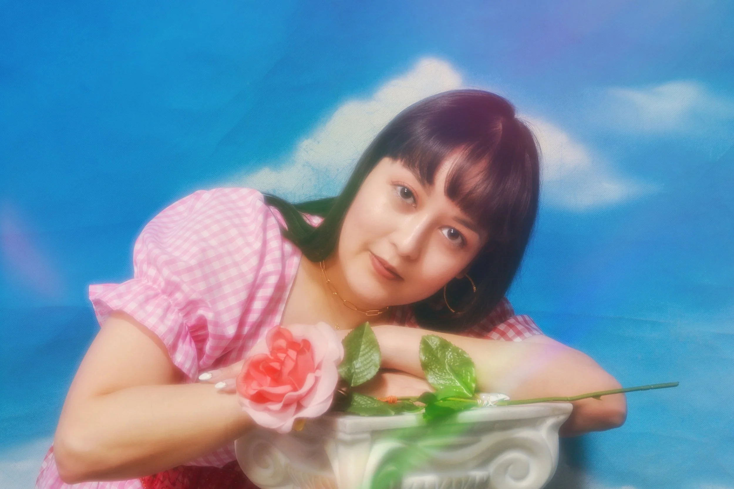 Young woman with dark hair and bangs, wearing a pink gingham top, resting her face on her folded arms on a decorative white pedestal with a pink rose and green leaves, against a blue sky with clouds background.