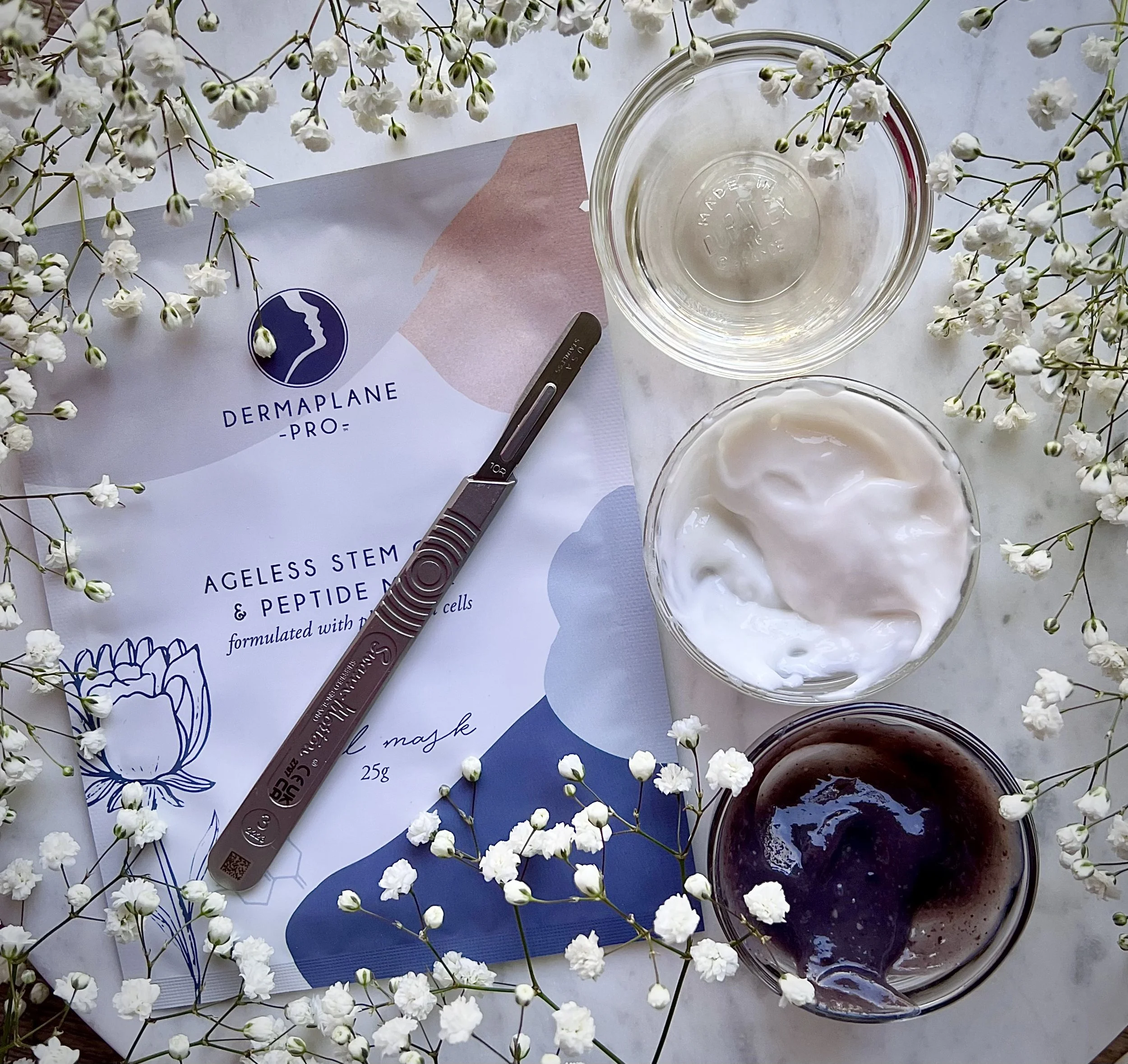 Beauty skincare products with a spatula, a box labeled Dermaplane Pro Ageless Stem and Peptide Mask, two glass bowls with a clear liquid, white cream, and dark gel, all surrounded by white baby's breath flowers.