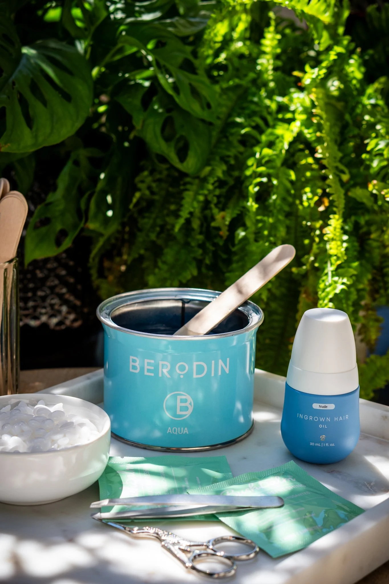A manicure setup outdoors with a blue Berodin Aqua wax container, a small bowl of white nail tips, a blue and white hair oil bottle labeled "Nude Ingrown Hair Oil," a pair of nail scissors, a nail file, and green nail wraps, all on a white surface with lush green plants in the background.