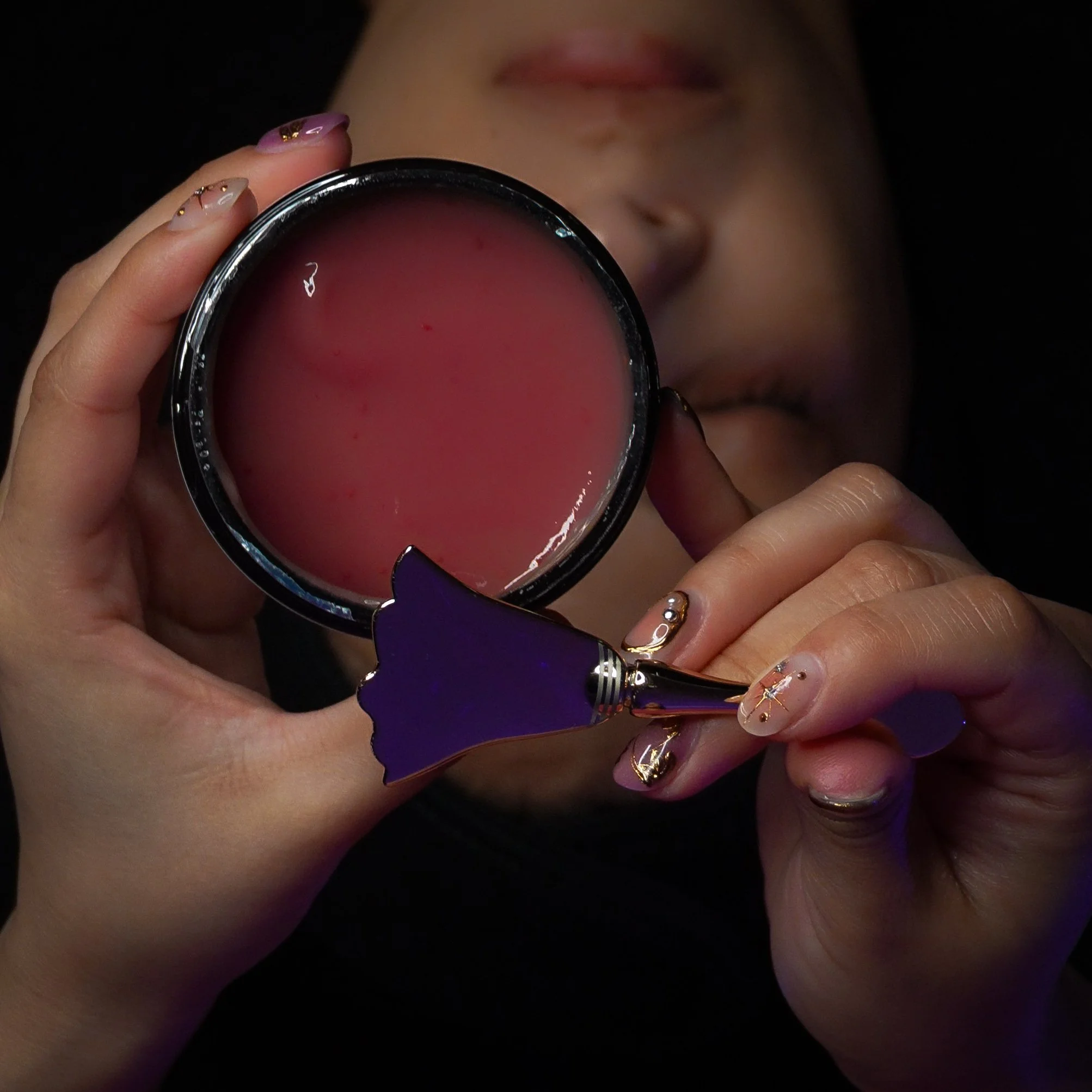Close-up of a woman holding skincare product  filled with a pink facial mask, with a gold fan-shaped spoon in her hand, and a face partially visible in the background.