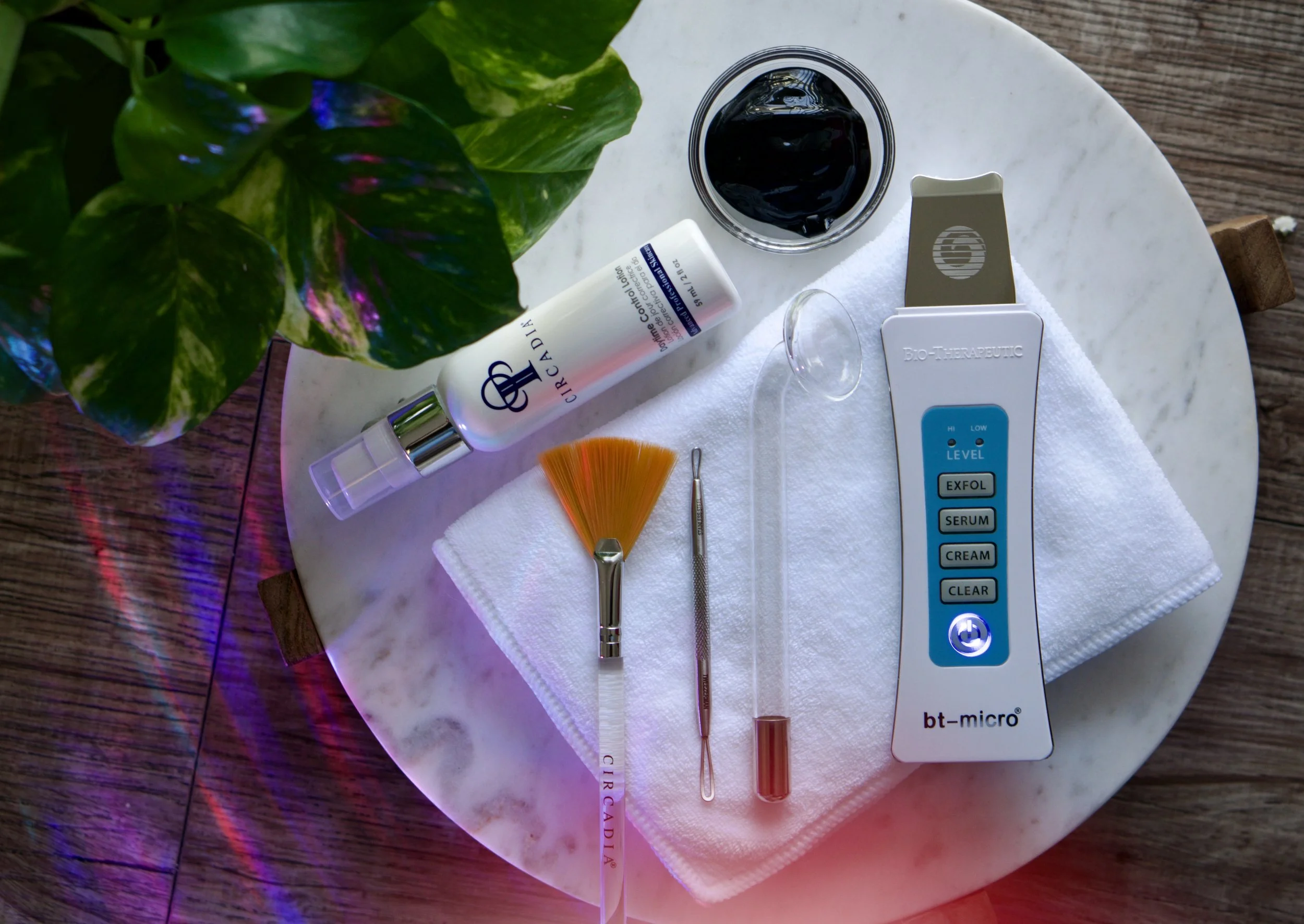 Skincare and beauty tools on a white marble table, including a bottle of serum, a glass jar with black gel, a white towel, a fan brush, a metal tool, a test tube, and a facial device with buttons labeled exfoliation, serum, cream, and clear.