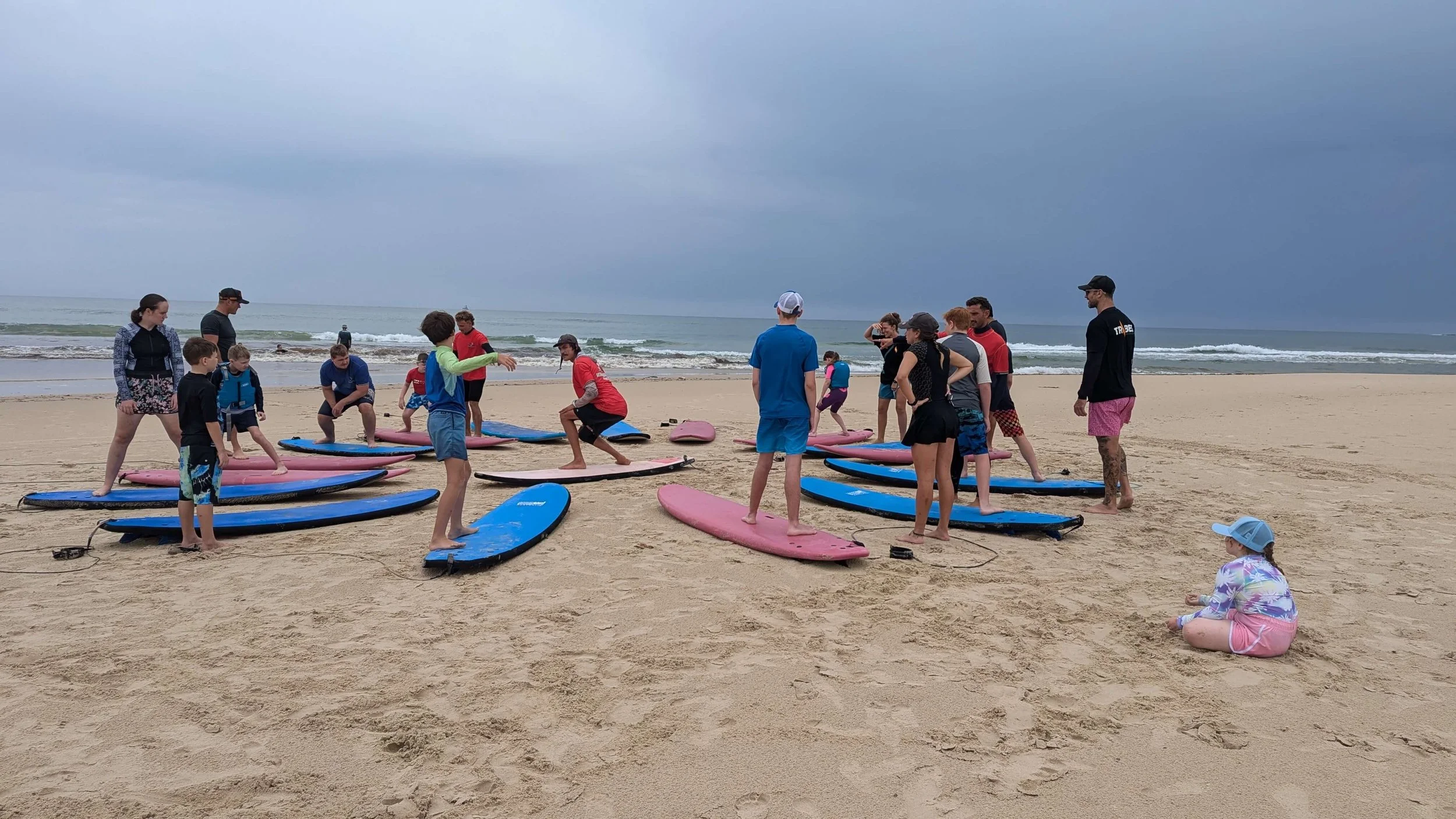 Group of people, including children and adults, on a sandy beach preparing for paddleboarding, with surfboards laid out on the sand, ocean waves in the background, and cloudy sky overhead.
