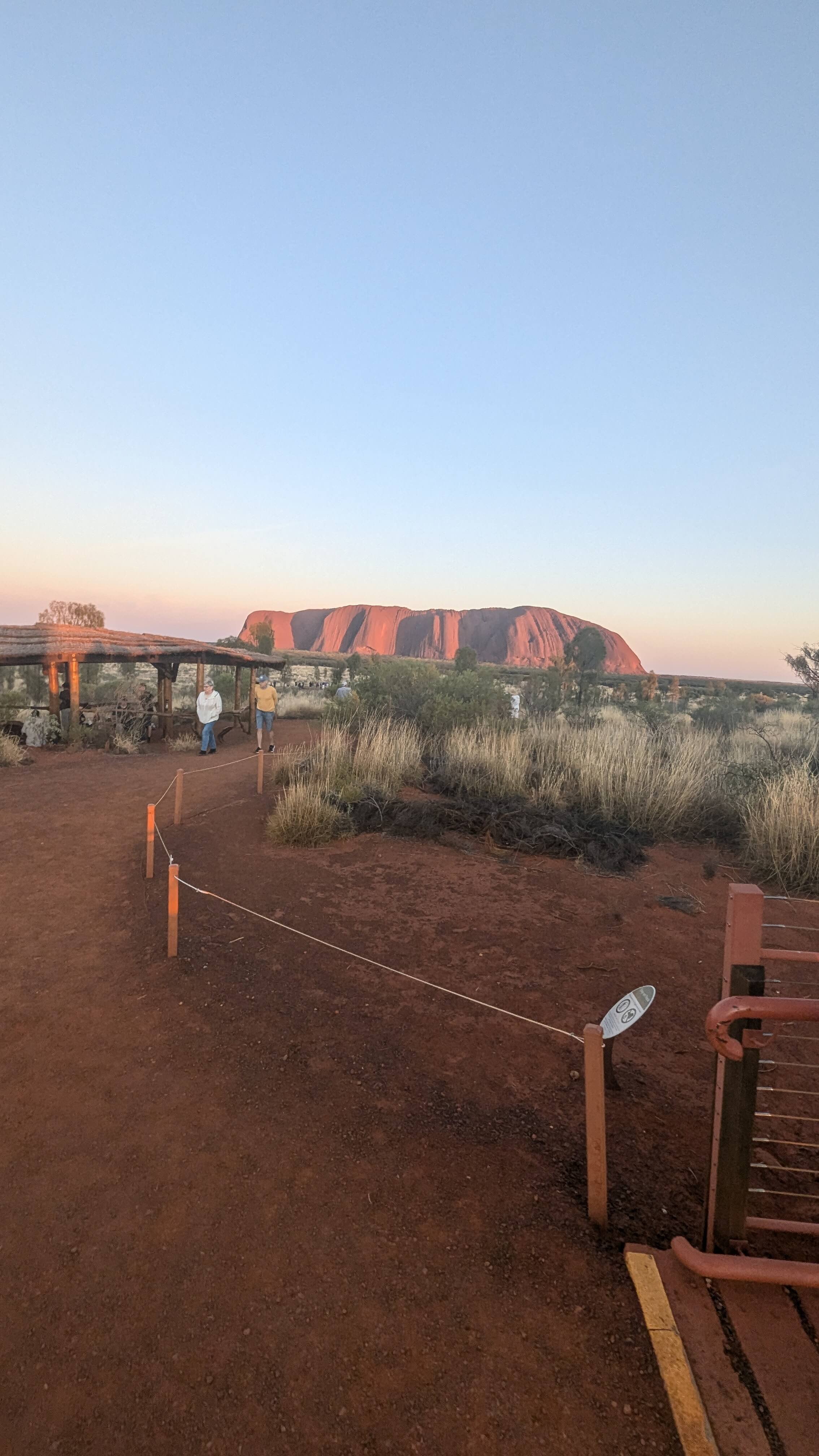 View of Uluru (Ayers Rock) in the Australian desert at sunset, with a few people walking along a dirt path, some greenery, and a small shelter in the foreground.