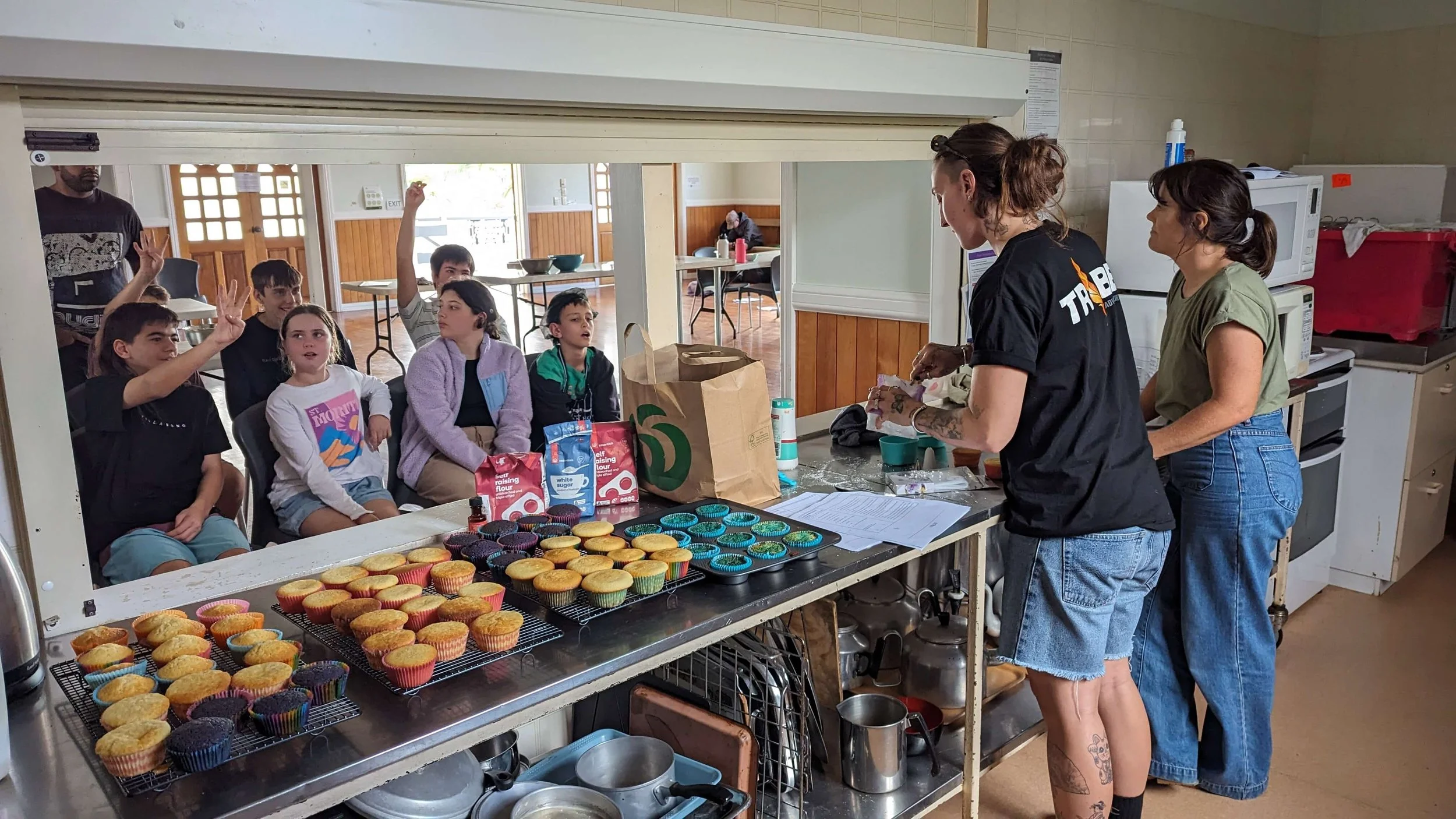 A group of children sitting and raising their hands in a cafeteria or community kitchen, with two women preparing baked goods at a counter with cupcakes.