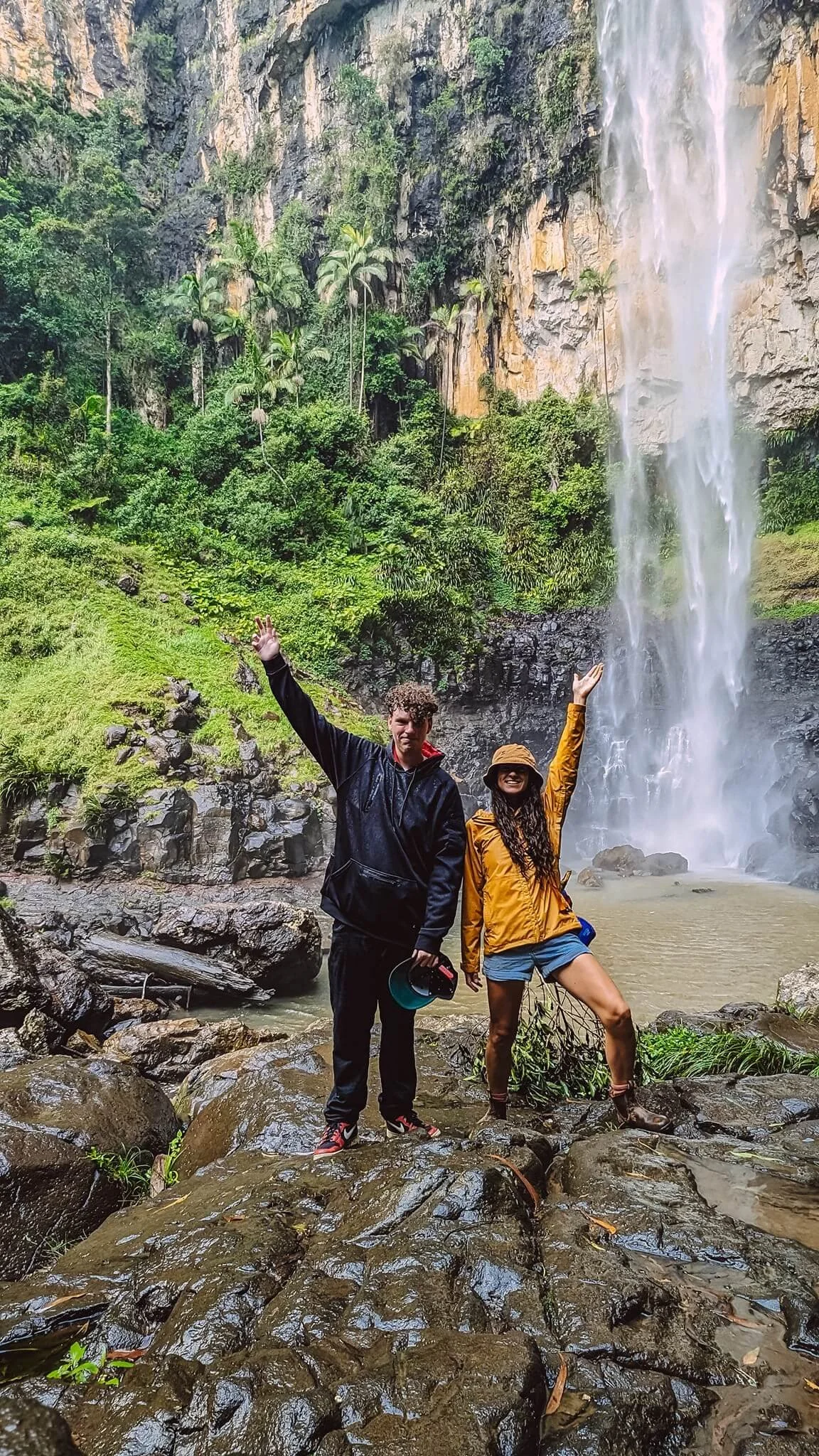 Two people standing on wet rocks in front of a waterfall and lush green forest, with one person raising their hand and the other posing with one leg up.