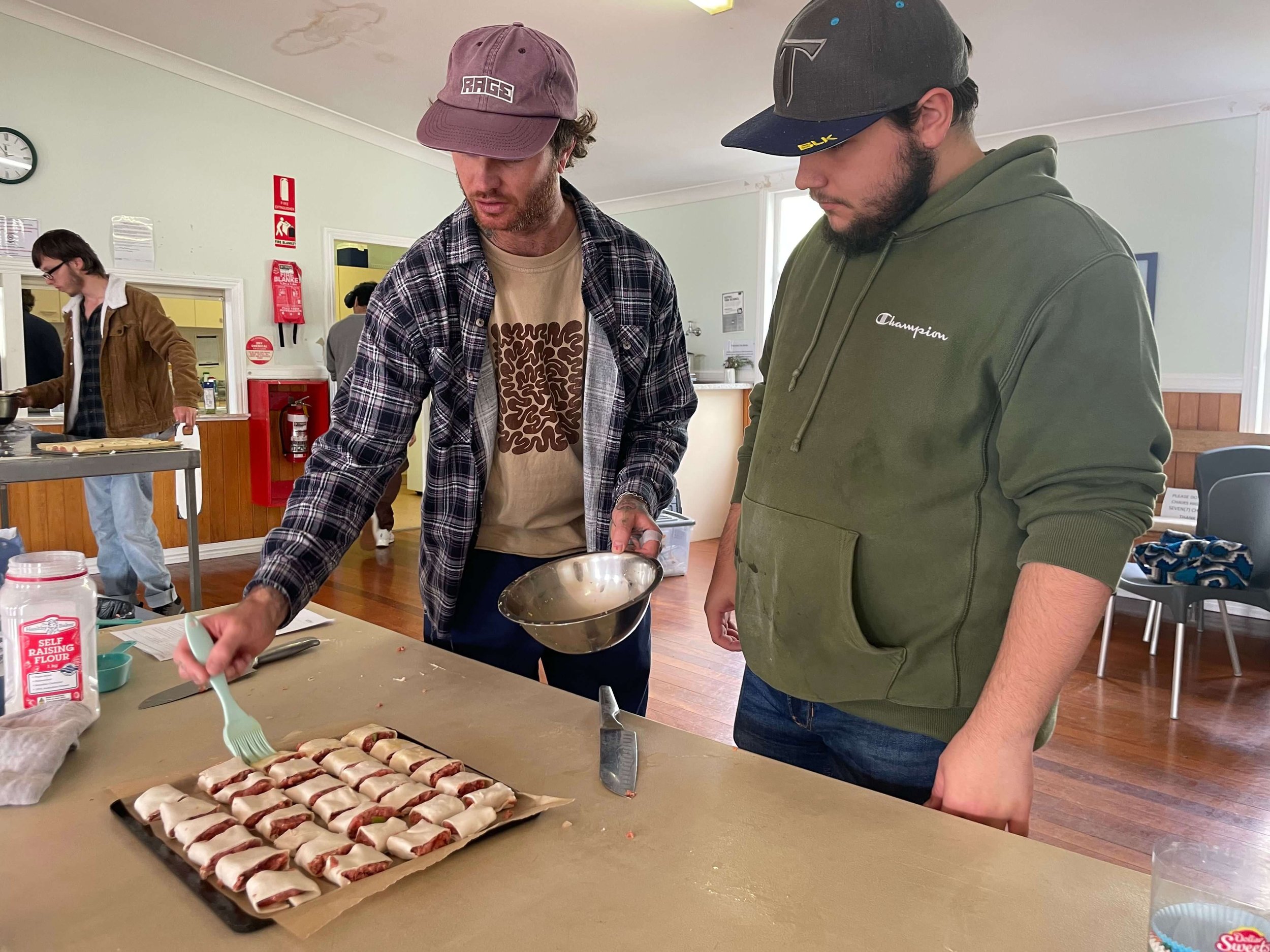 Two men preparing food at a kitchen table with sausage rolls, one pouring sauce and the other watching. In the background, a woman is cooking.
