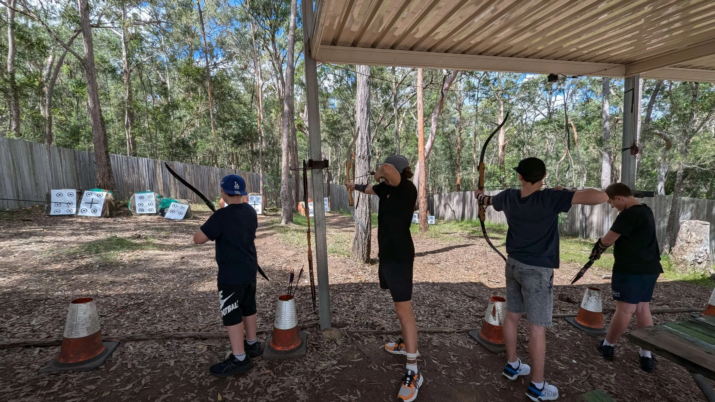 Four boys and a woman practicing archery under a covered outdoor area, with targets set up on a wooded background.