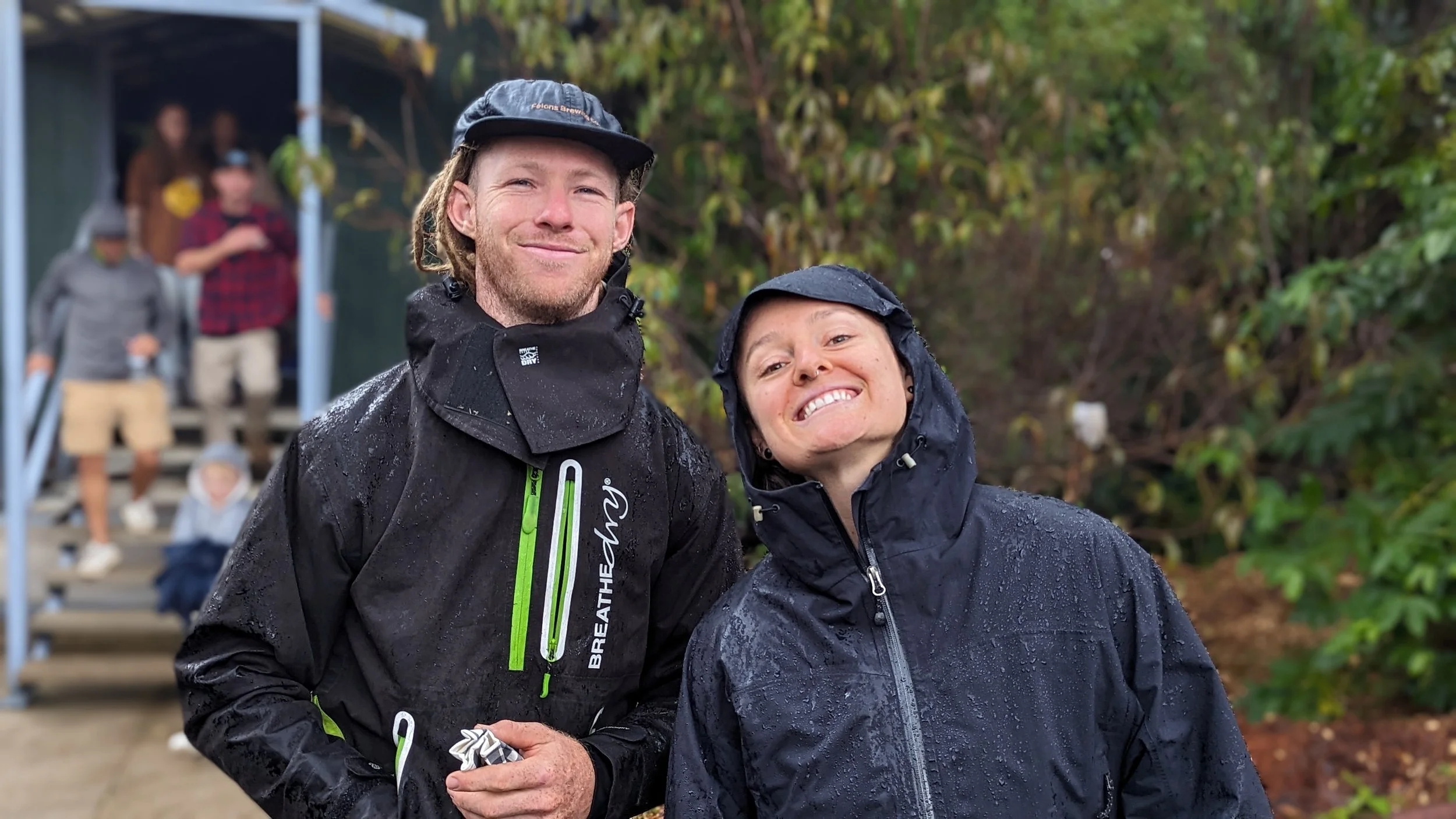 A man and woman smiling and posing outdoors in rainy weather, wearing black waterproof jackets and hats, with a background of trees and a group of people on stairs.