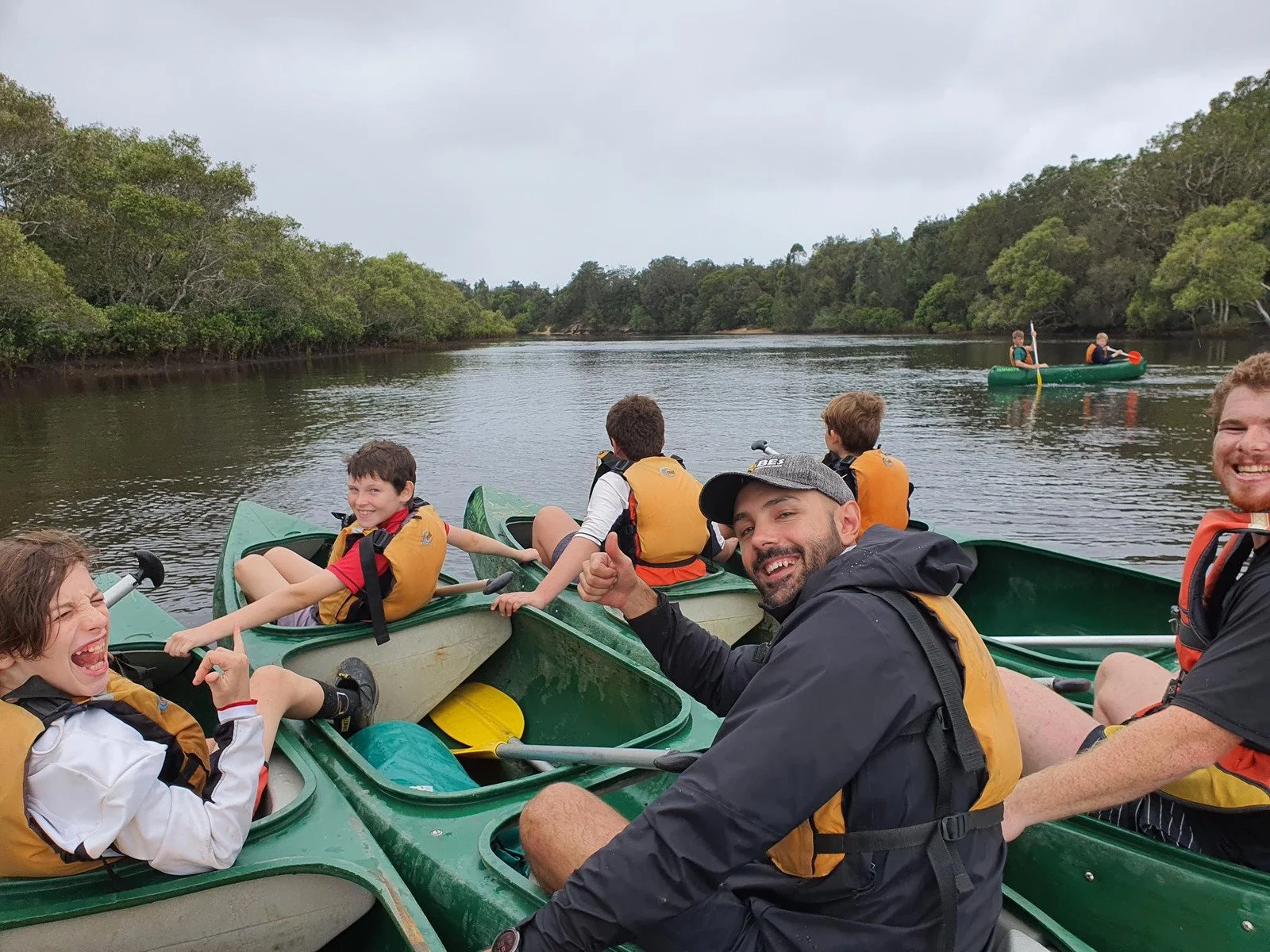 Group of kids and adults in kayaks on a calm river surrounded by trees, enjoying kayaking.