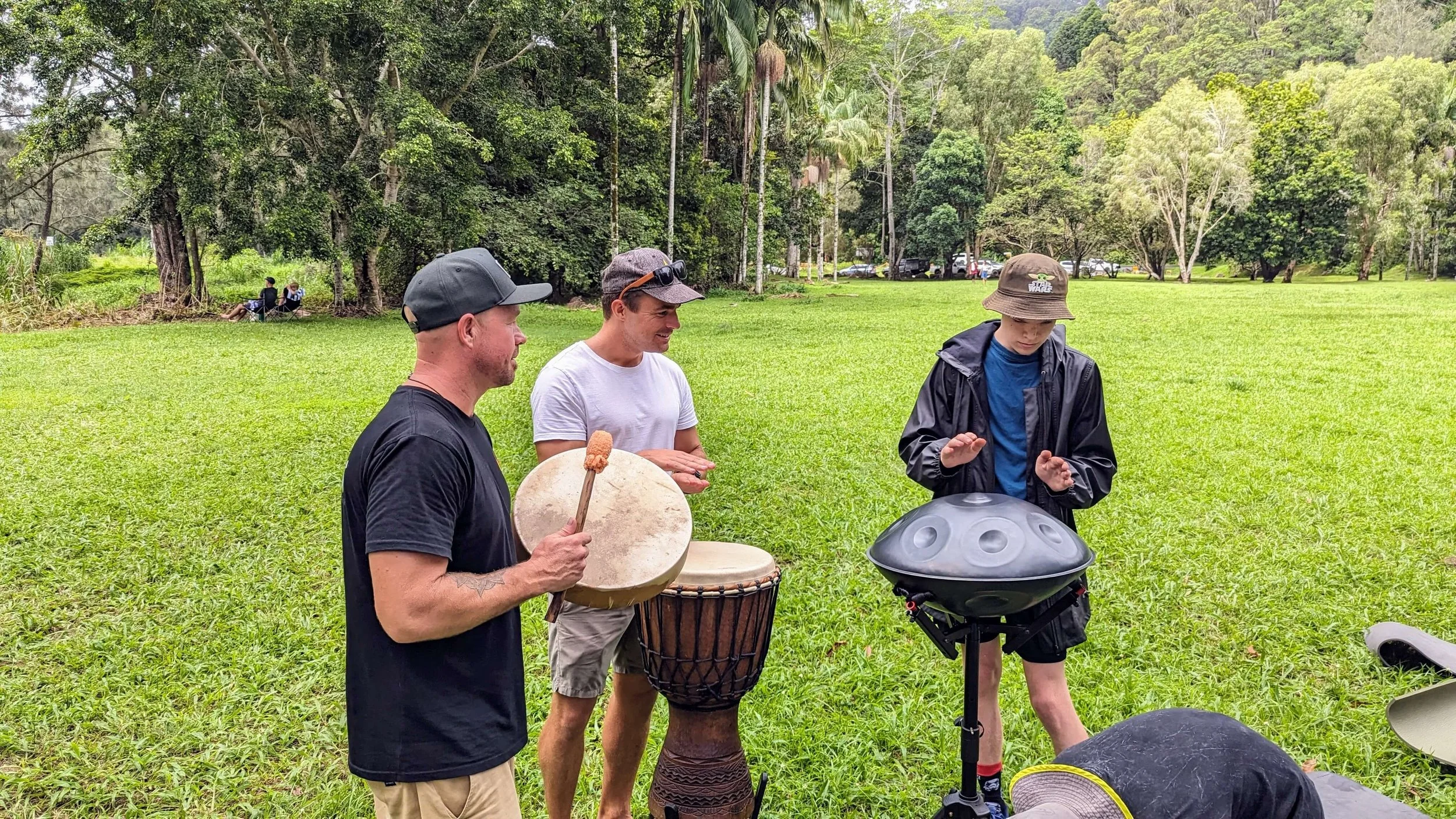 Three men standing in a grassy park playing musical instruments. One is holding a drumstick and a small drum, the second is playing a djembe drum, and the third is playing a steel tongue drum. They are surrounded by lush trees and greenery.