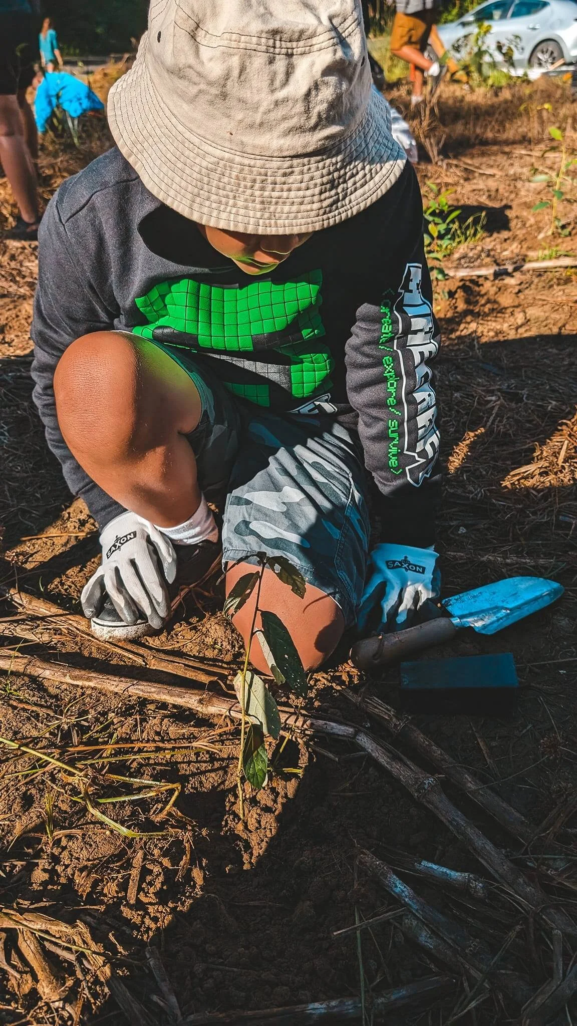 Child planting a seedling in bare soil, wearing a gray bucket hat, black jacket with green and white graphics, camouflage shorts, and gloves, with gardening tools nearby, and other people working in the background.